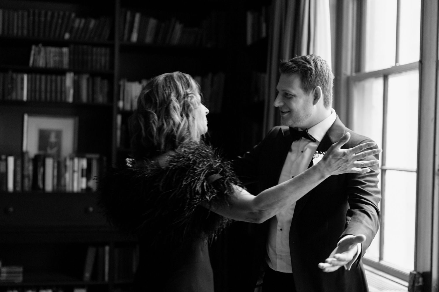 A couple dances intimately by a window in a library with bookshelves lining the walls in black and white.