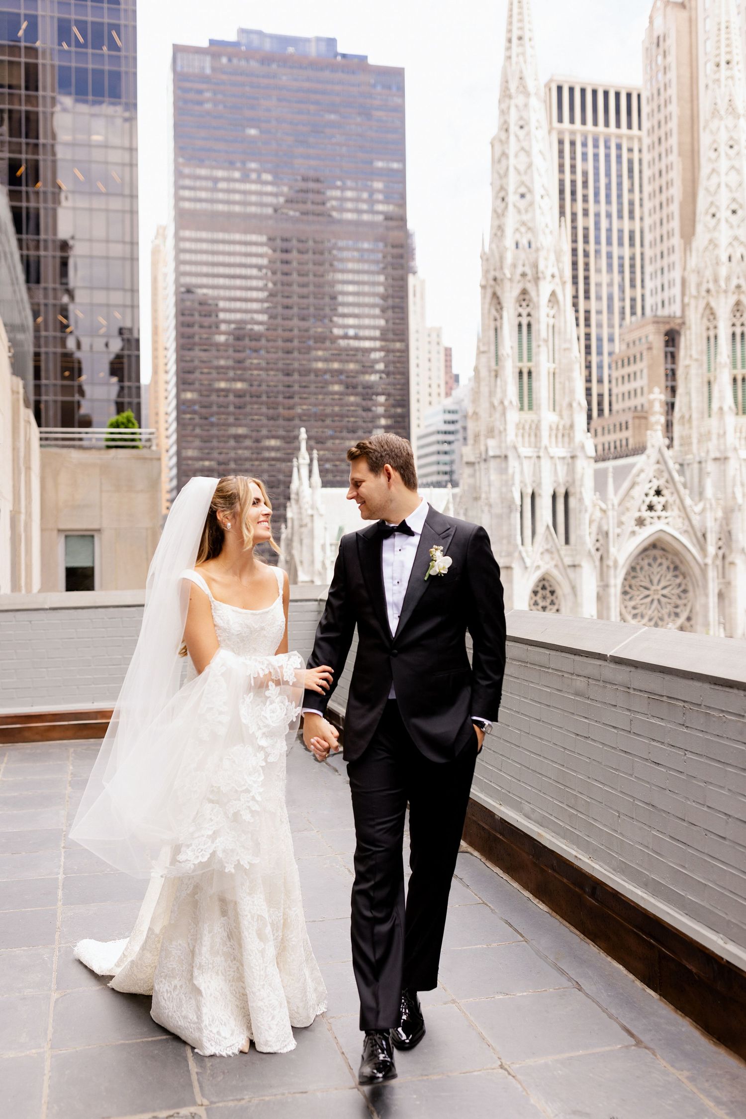 A couple in wedding attire walks along a rooftop with St. Patrick's Cathedral in New York City background.