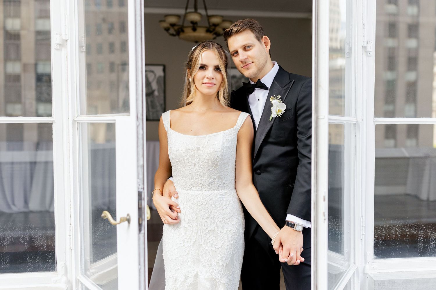 A newlywed couple poses together in formal wedding attire in a bright doorway setting.