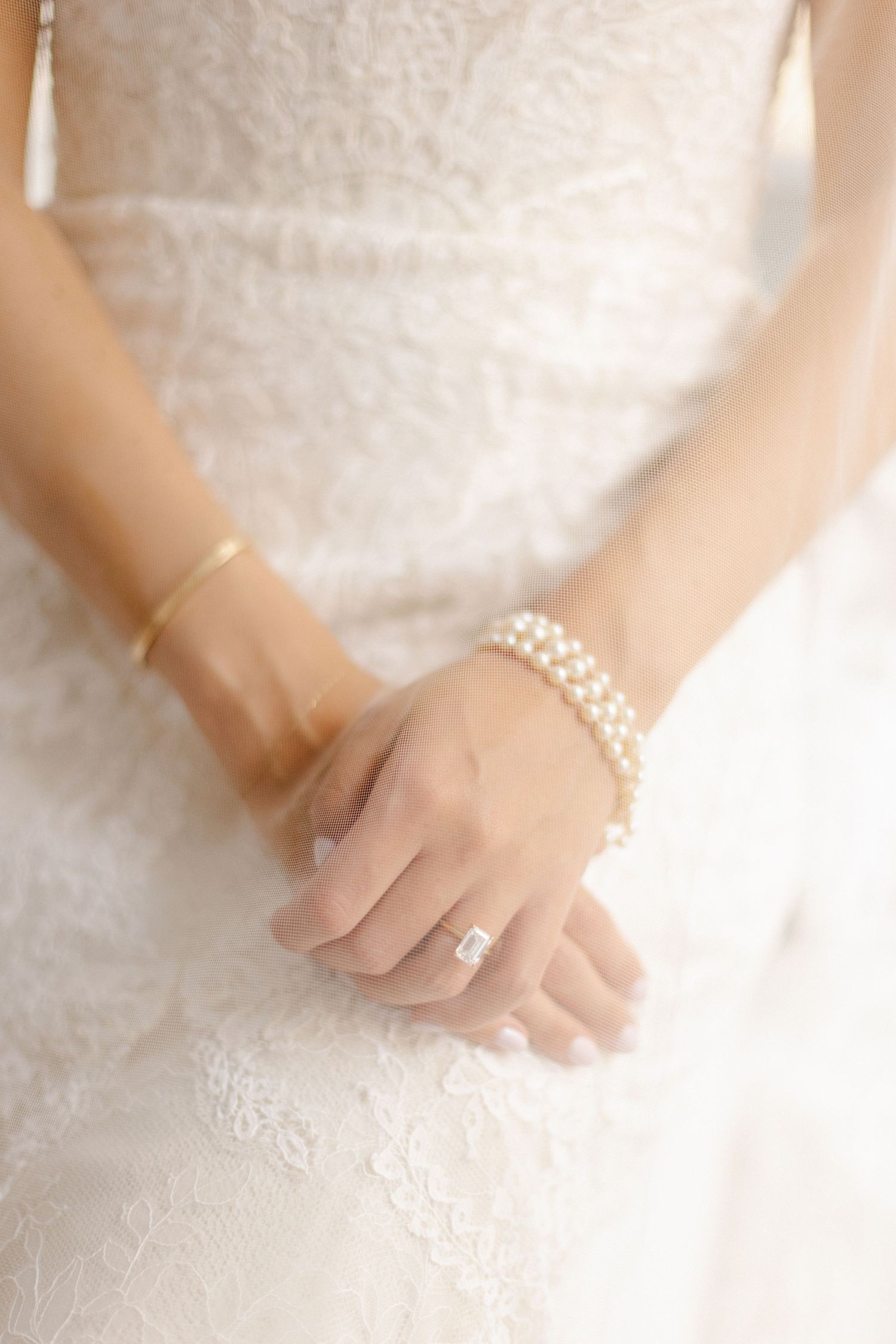 Close-up of bride's hands showcasing wedding ring and pearl bracelet against white lace dress.