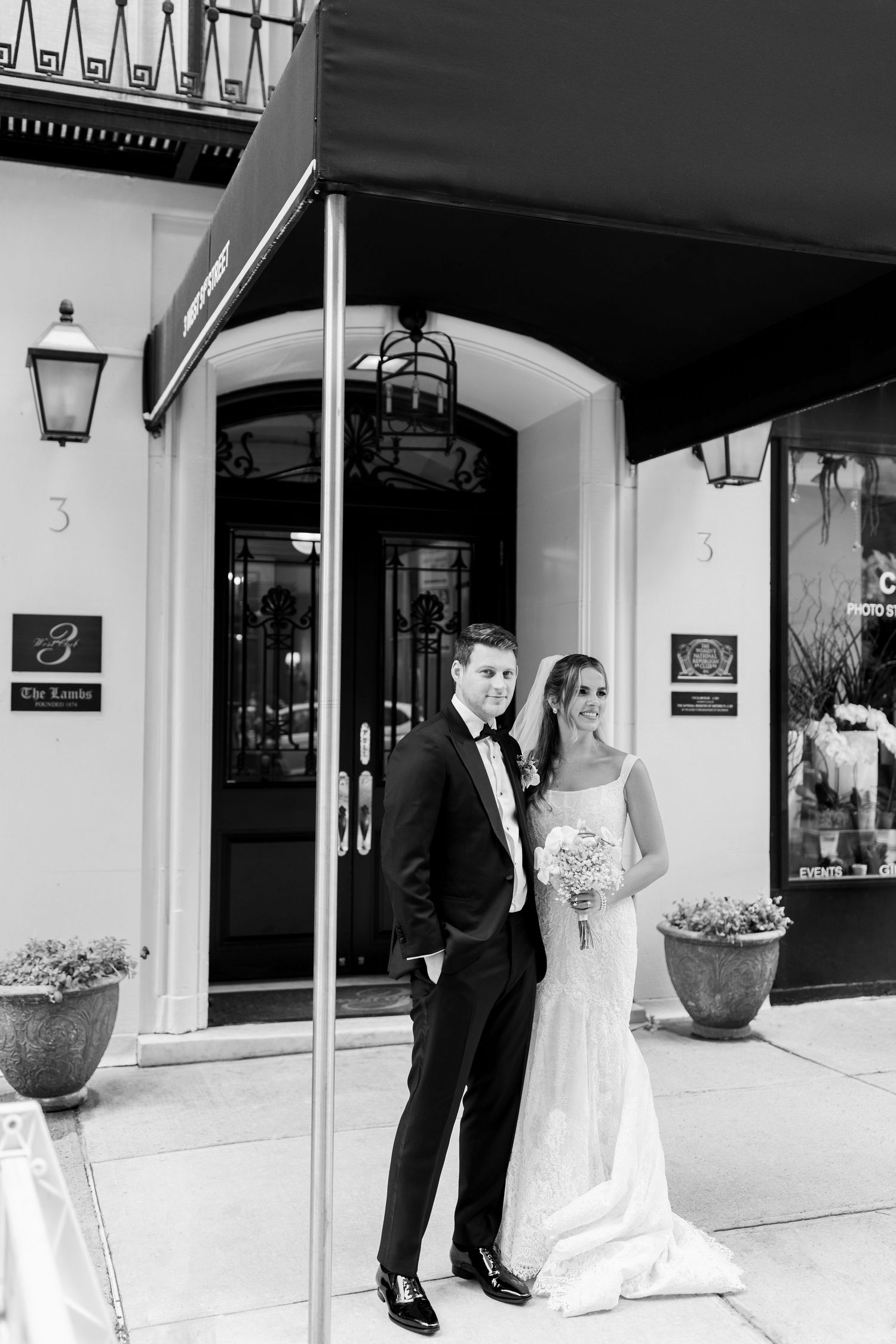 A formal couple posing in front of an elegant building entrance with potted plants and decorative lanterns.