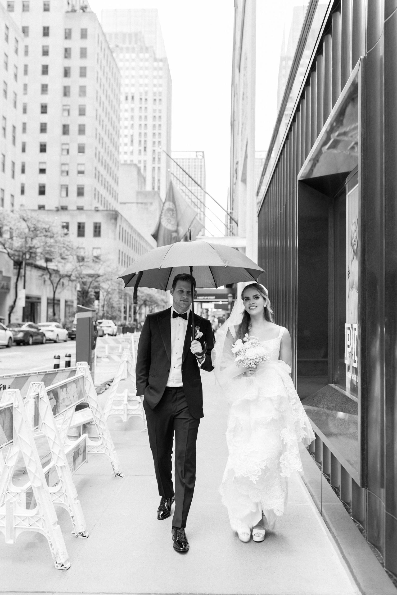 A pair walks under an umbrella on a rainy city sidewalk in wedding attire.
