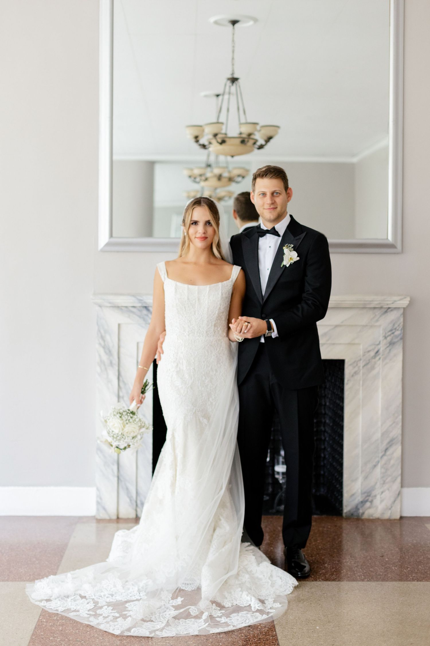 Elegant couple poses in formal wedding attire in front of marble fireplace.