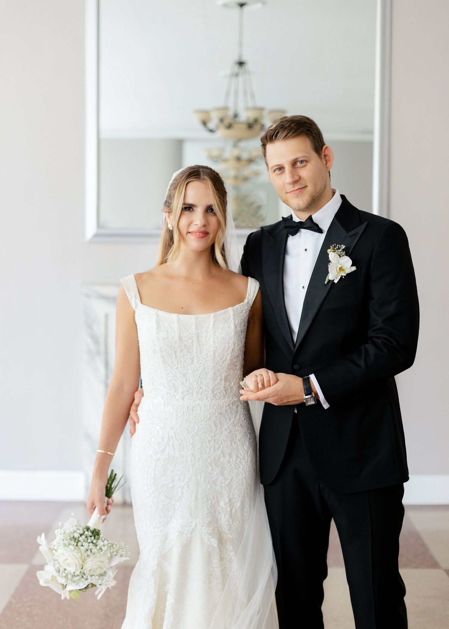 Elegant wedding couple in classic wedding attire pose together in a bright white room.