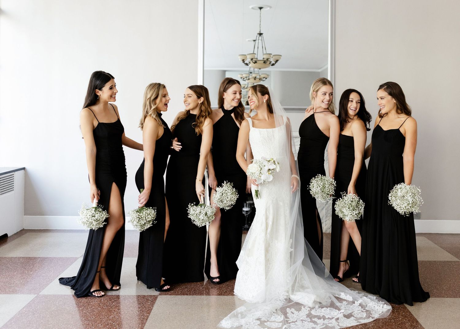 Elegant bridal party poses in a bright room with bridesmaids in black dresses holding white bouquets.
