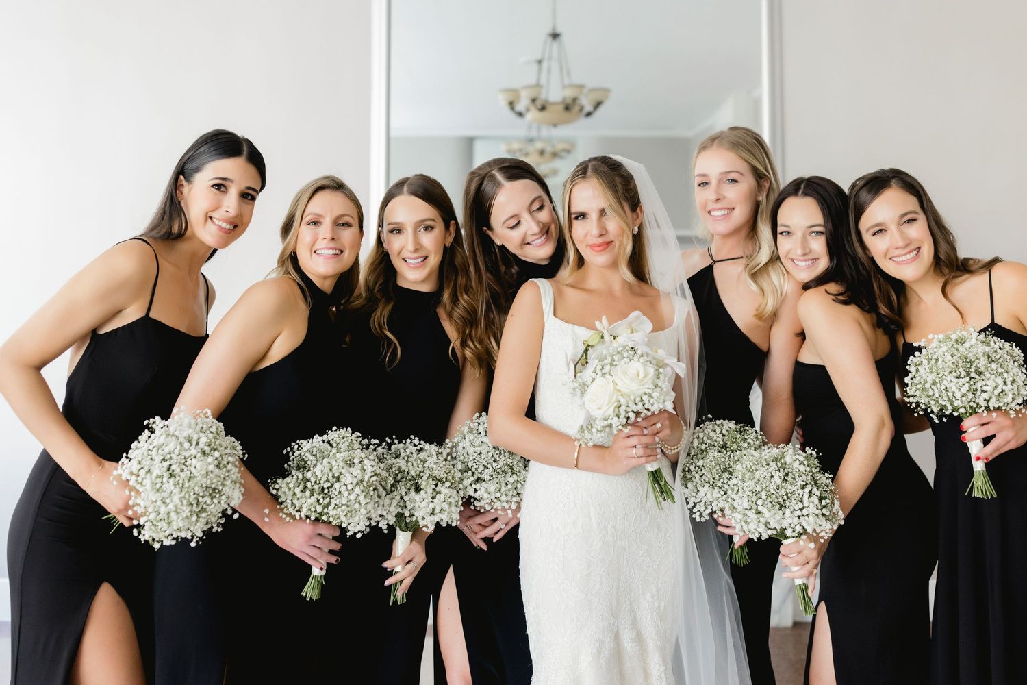 Bridal party in black dresses poses with the bride, all holding baby's breath bouquets.