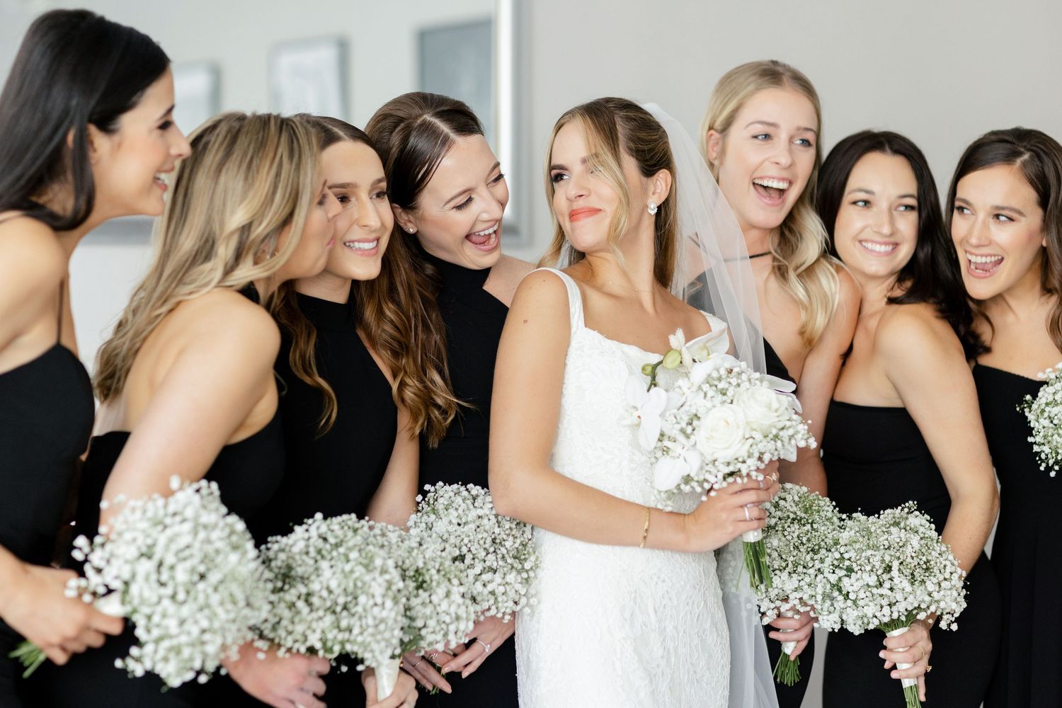 Wedding party in black bridesmaid dresses with white baby's breath bouquets laugh together with bride in white.