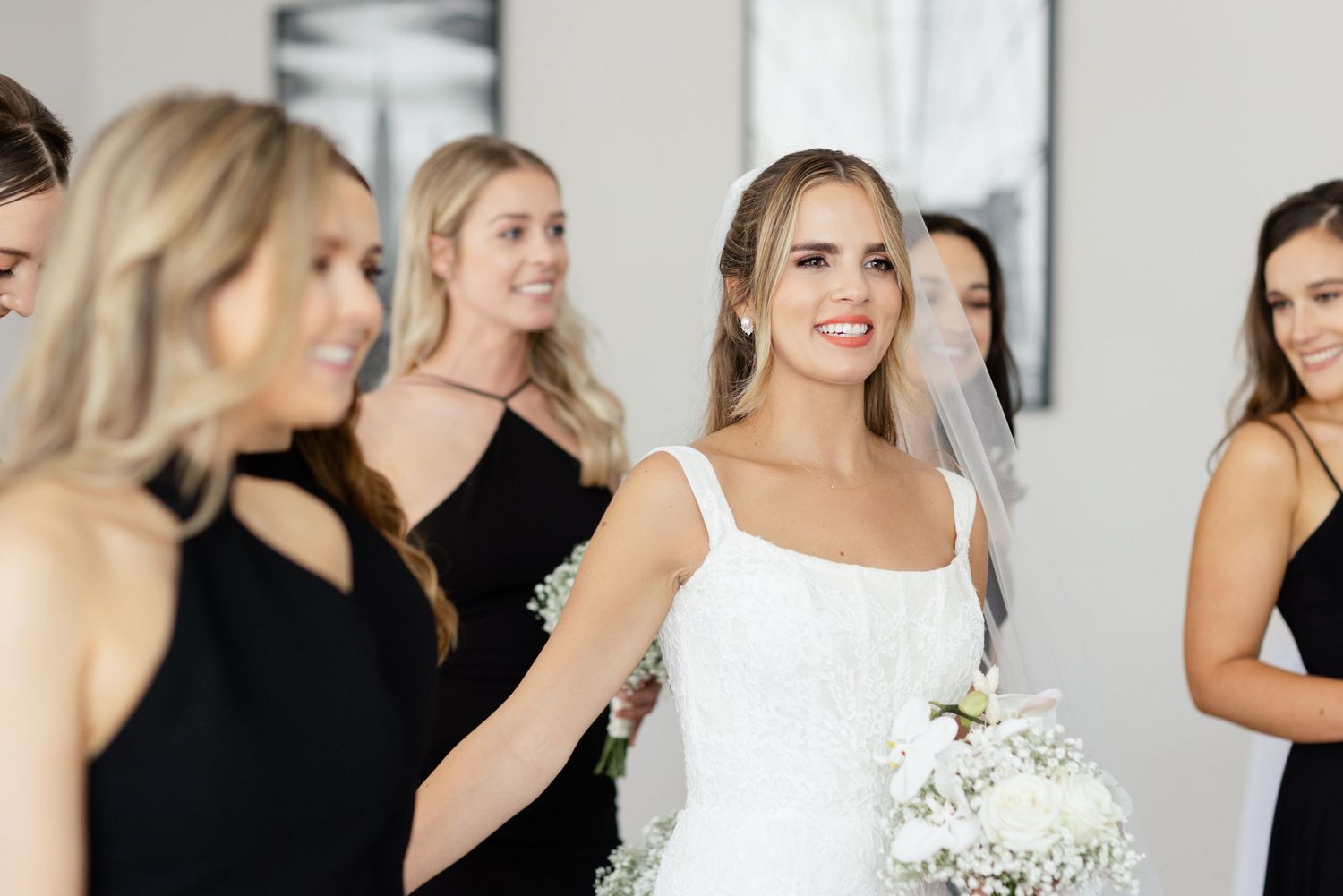 Bride in white dress smiles with bridesmaids wearing black dresses during wedding celebration.