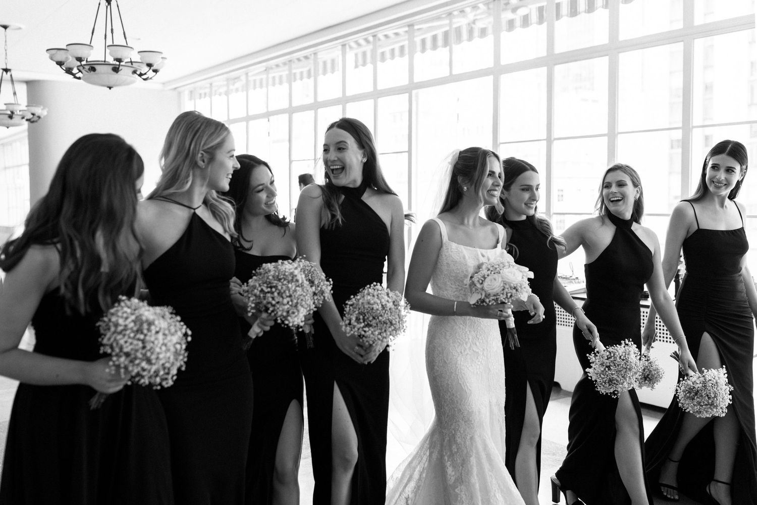 Wedding party members in black dresses gather with bouquets in bright sunlit room.