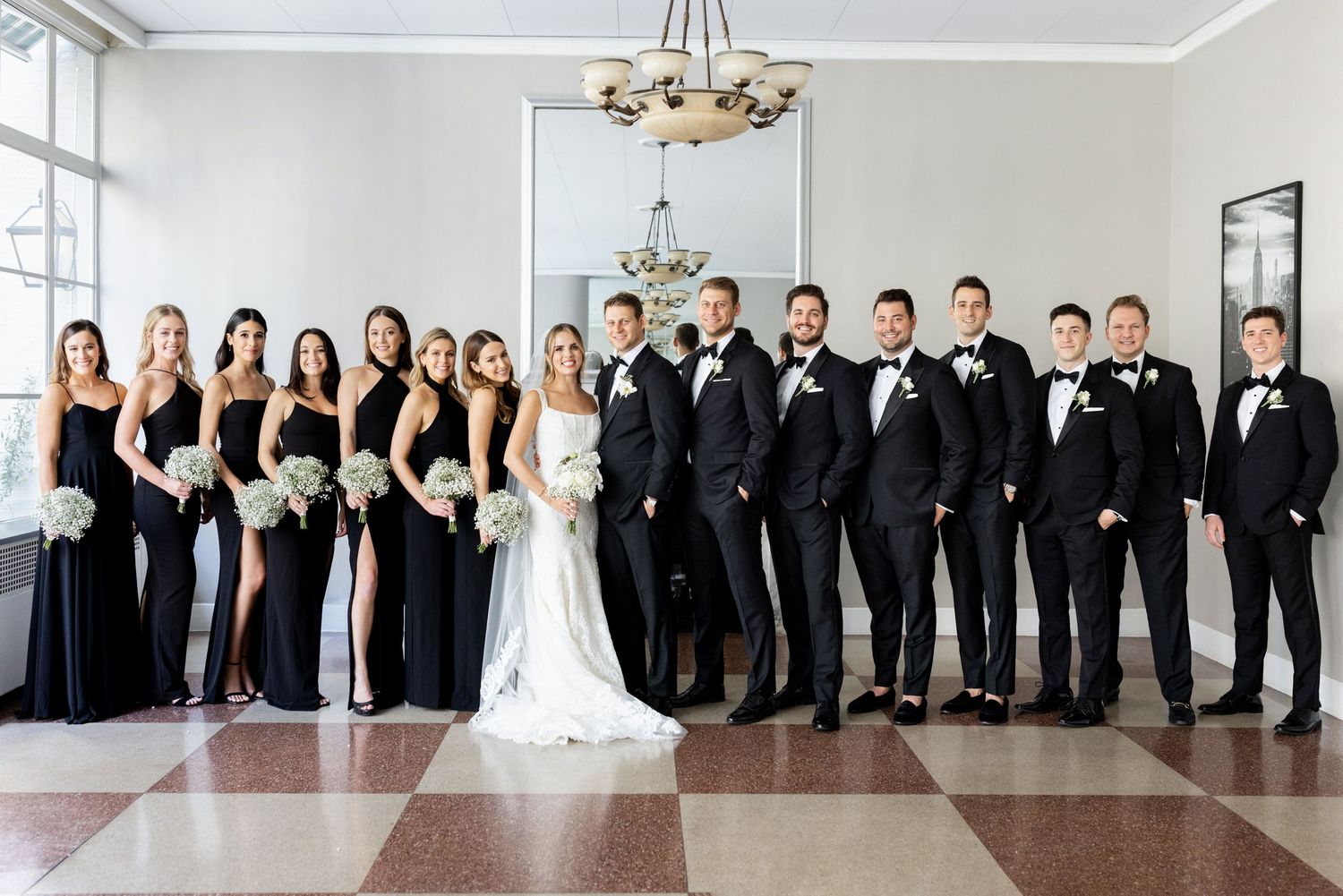 Wedding party in black and white attire poses formally in an elegant indoor setting with chandelier.