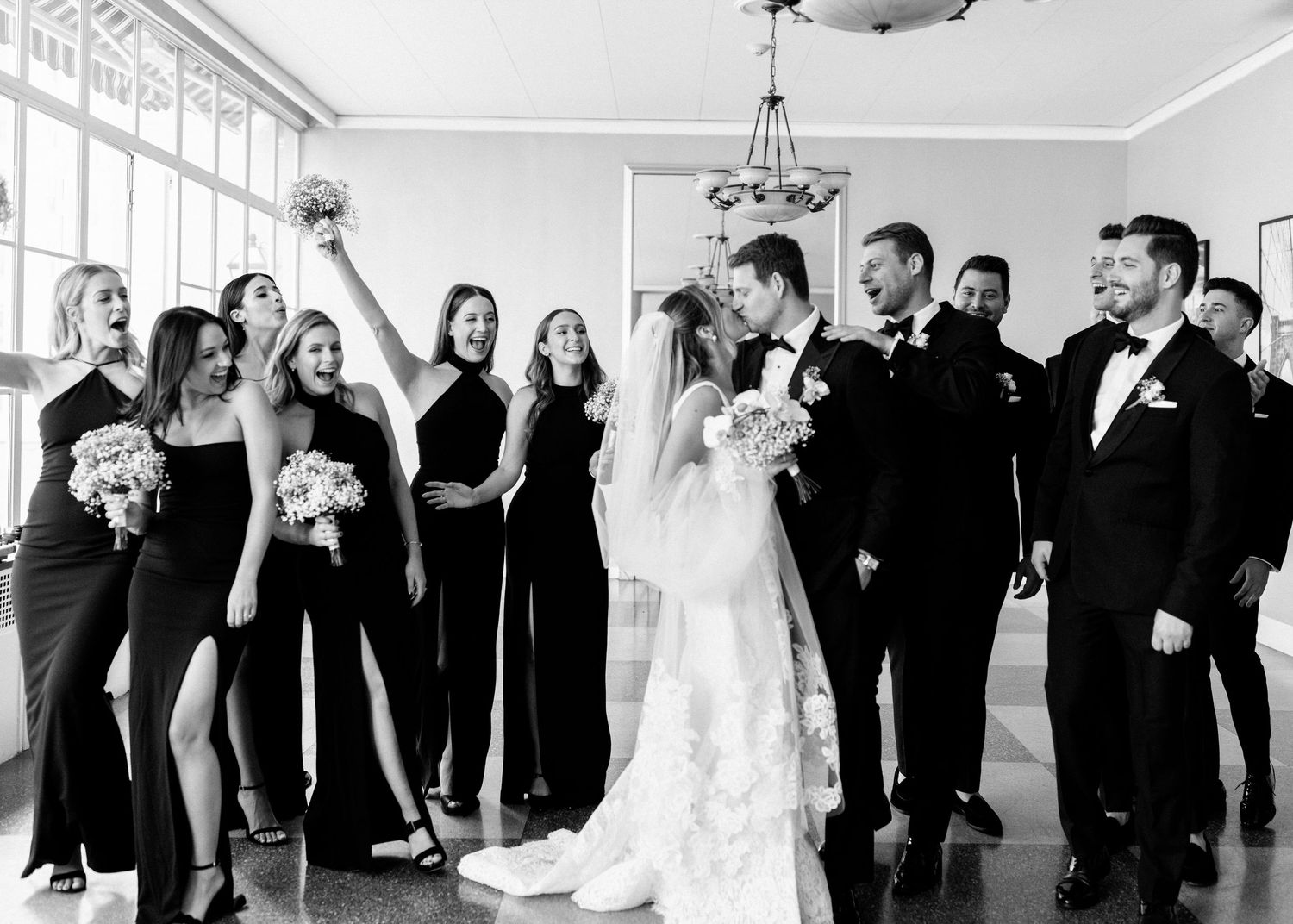 Wedding party in black attire celebrates with raised arms in bright indoor venue.