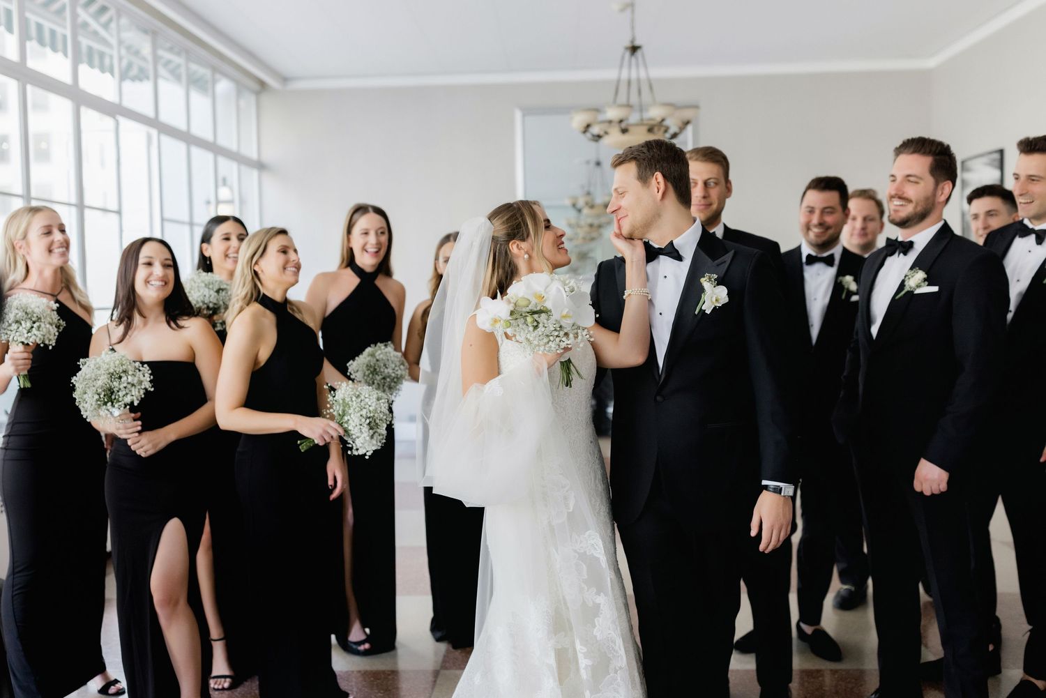 Wedding party in elegant black dresses and tuxedos pose together in a bright interior space.