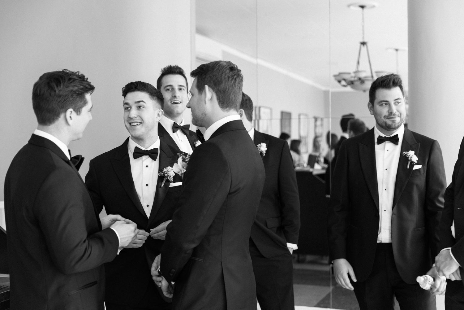 Groomsmen in formal black tuxedos gather for candid conversation at an elegant wedding venue.