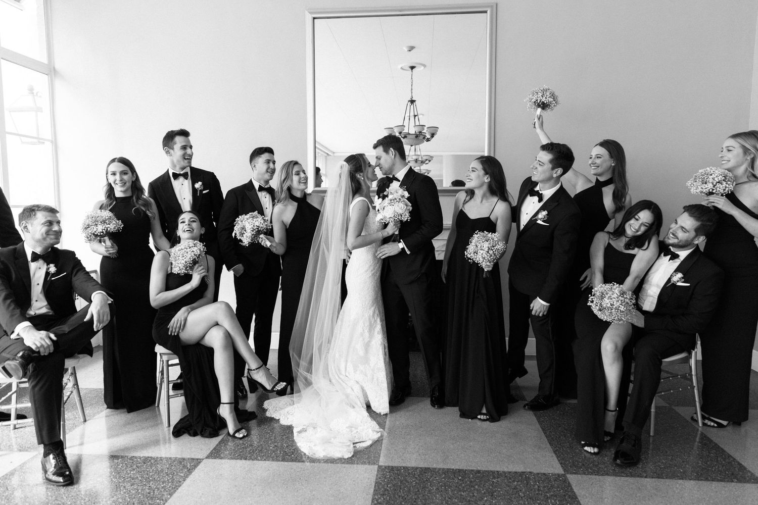 Wedding party in formal black attire poses for group photo in bright indoor setting.