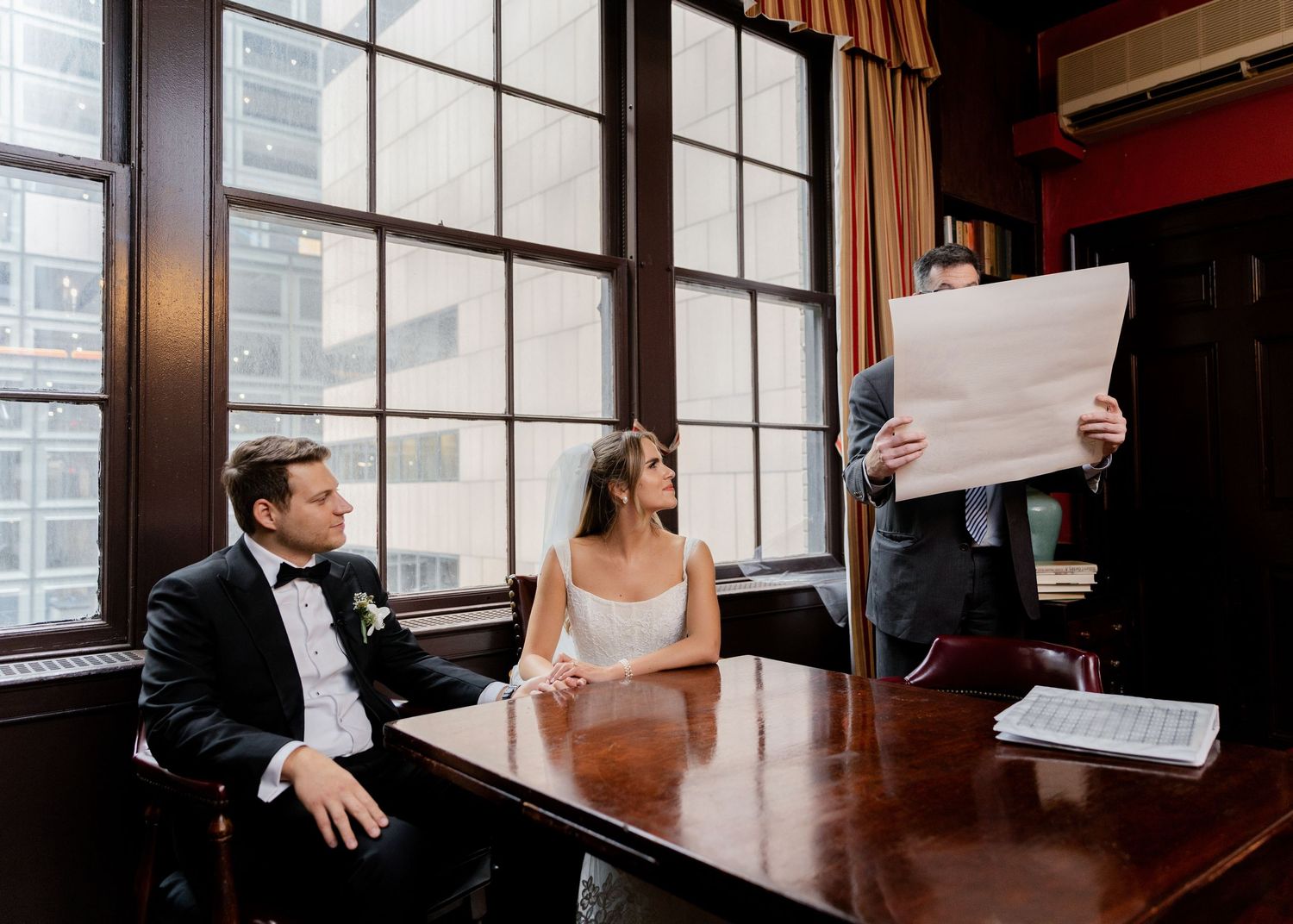Wedding couple sitting at table while reviewing documents in an elegant room with large windows.
