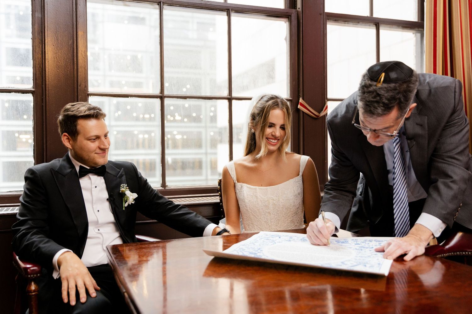 Wedding couple signing marriage documents with officiant at wooden table by window.