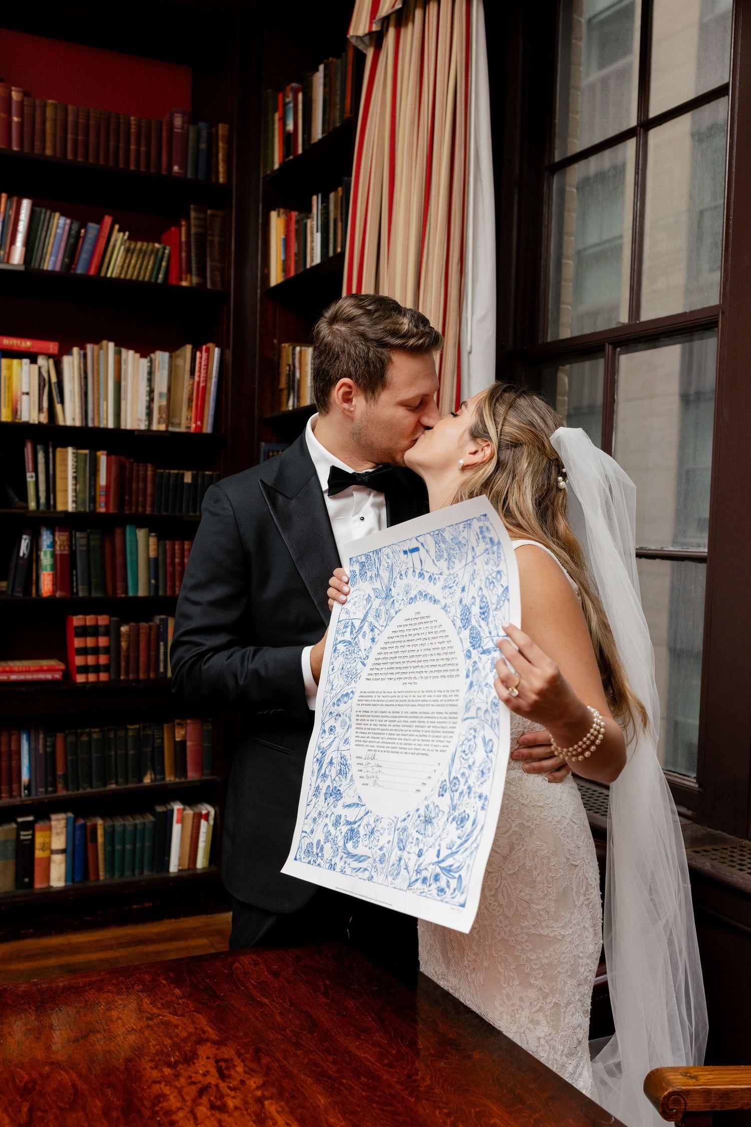 Romantic couple kissing in library while holding decorative certificate against bookshelf backdrop.