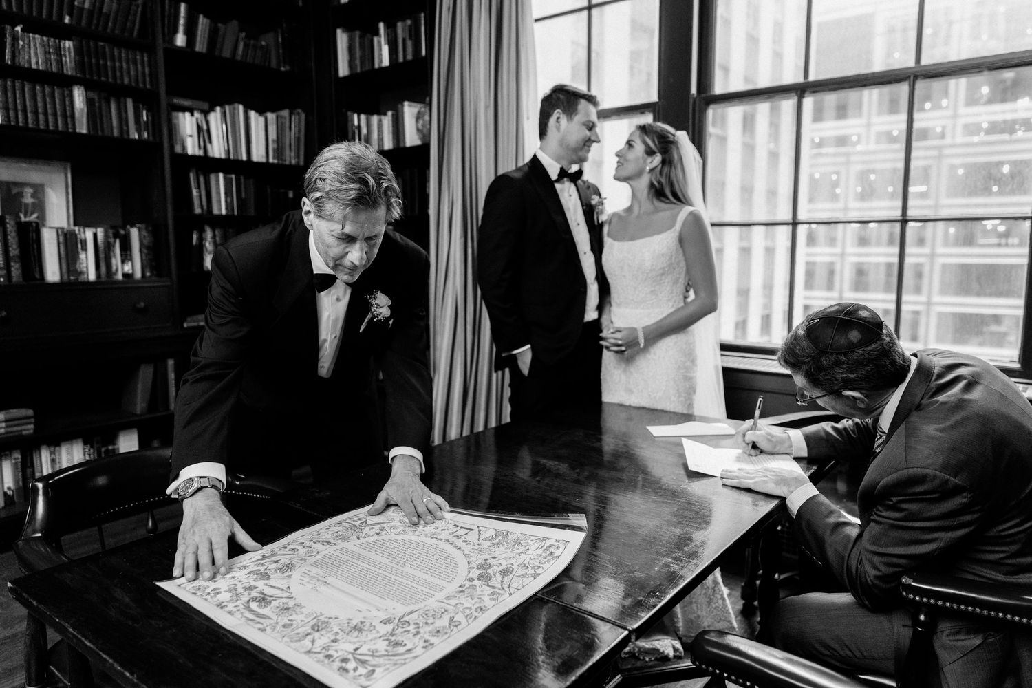 Wedding party gathered in a library examining documents on a desk in black and white.