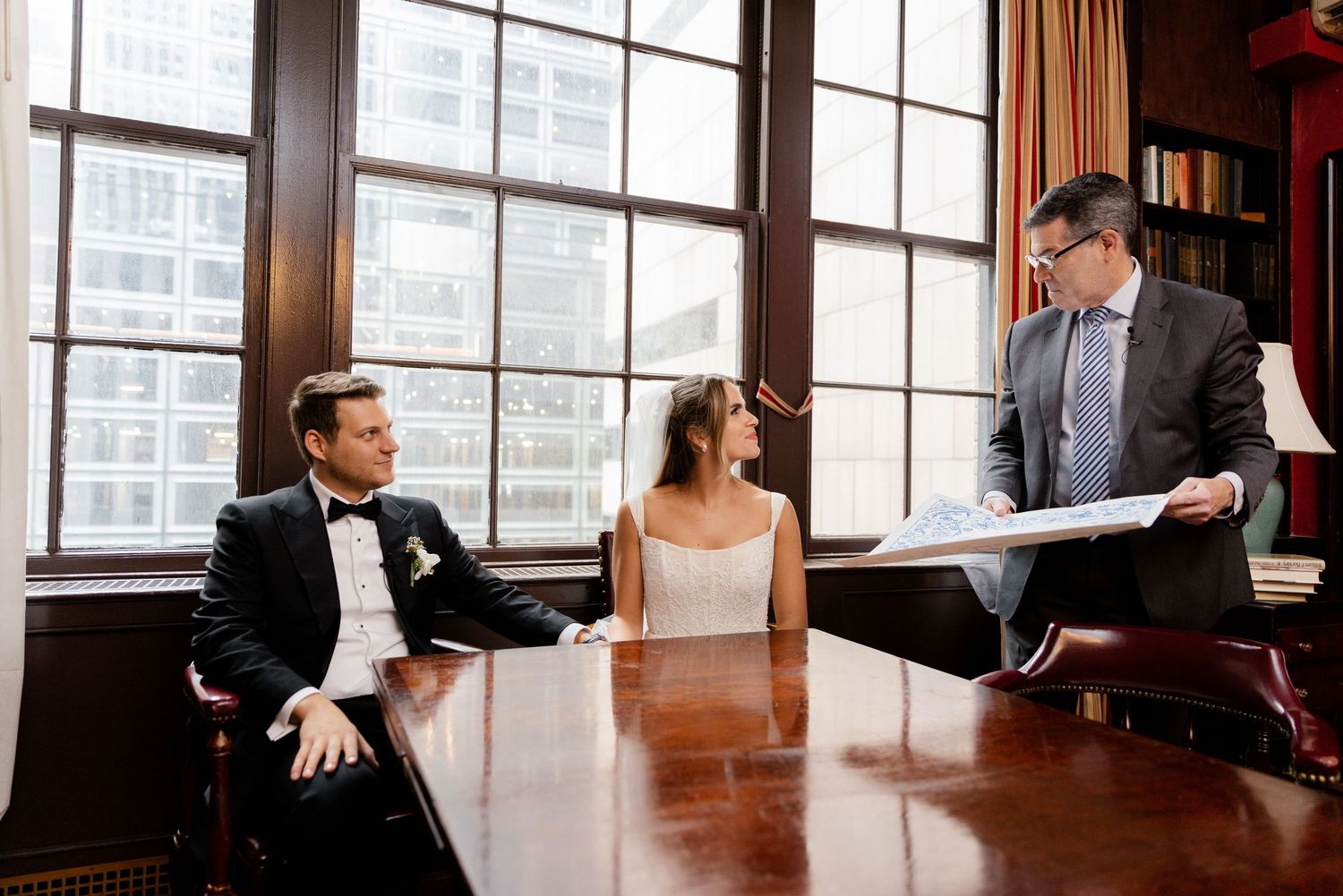 Three people in formal attire meeting at a wood-paneled office with large windows overlooking a city view.