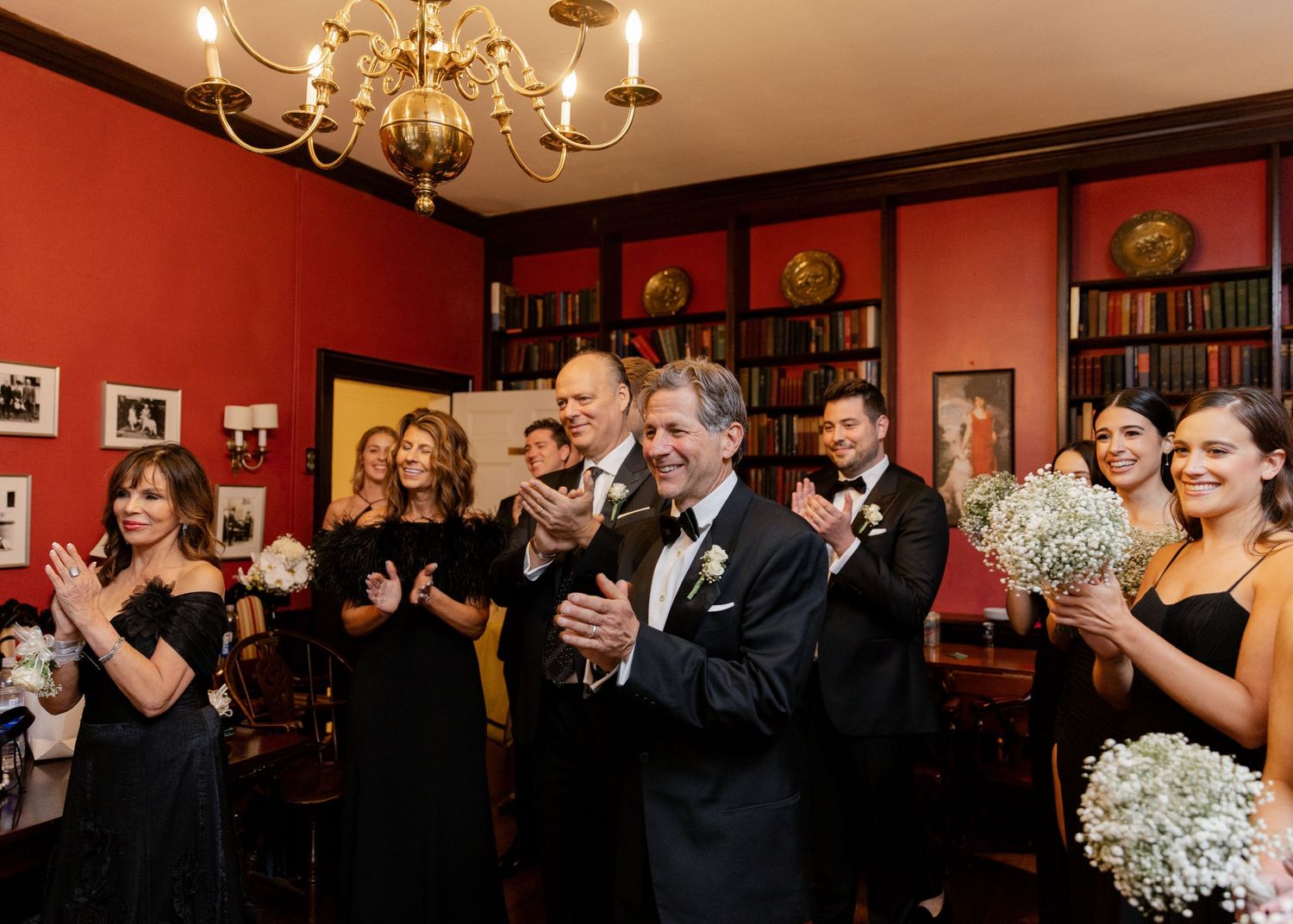 Wedding guests in formal attire applaud during celebration in a red-walled room with chandelier.