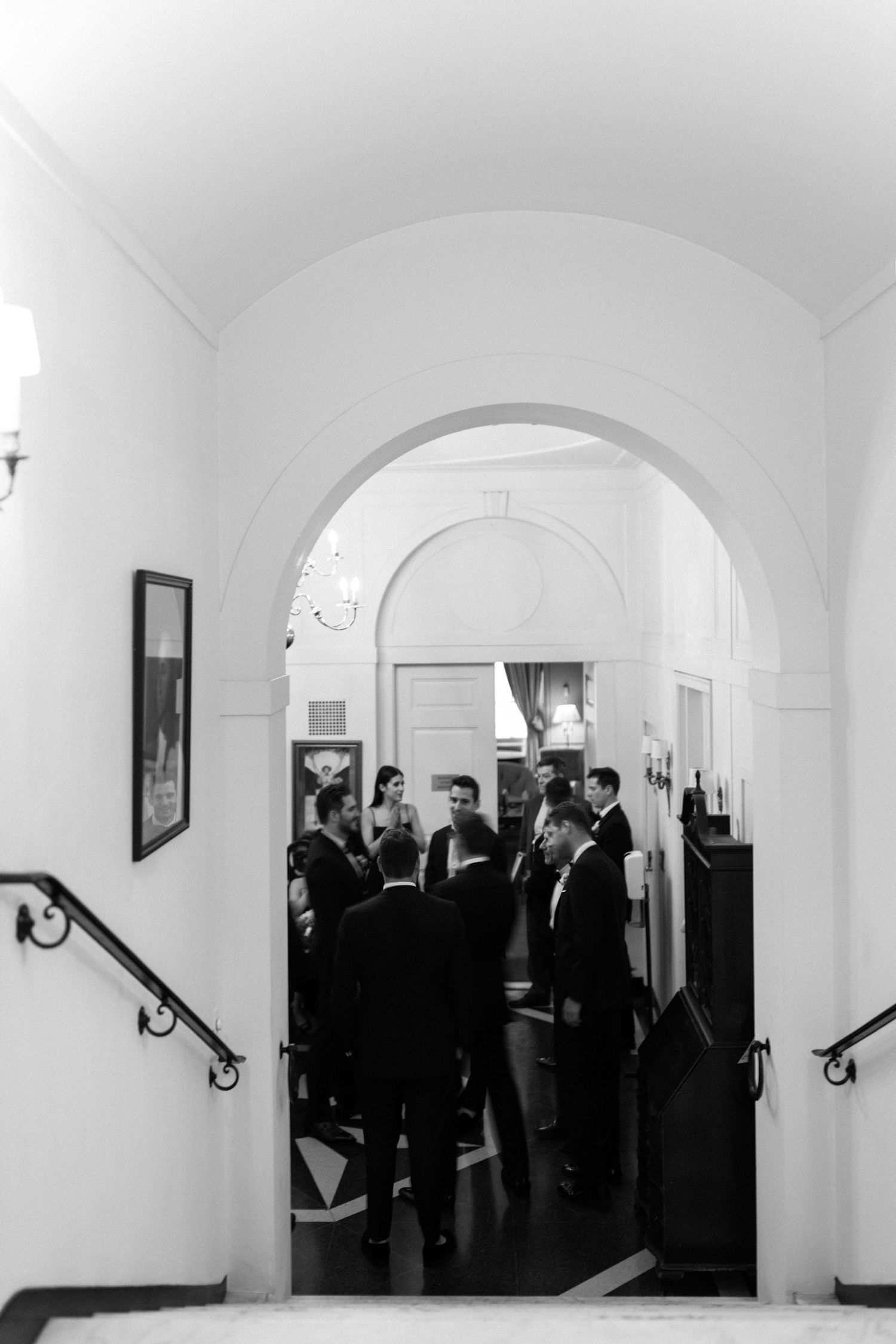 Black and white photo of wedding guests gathered under an elegant arched hallway.