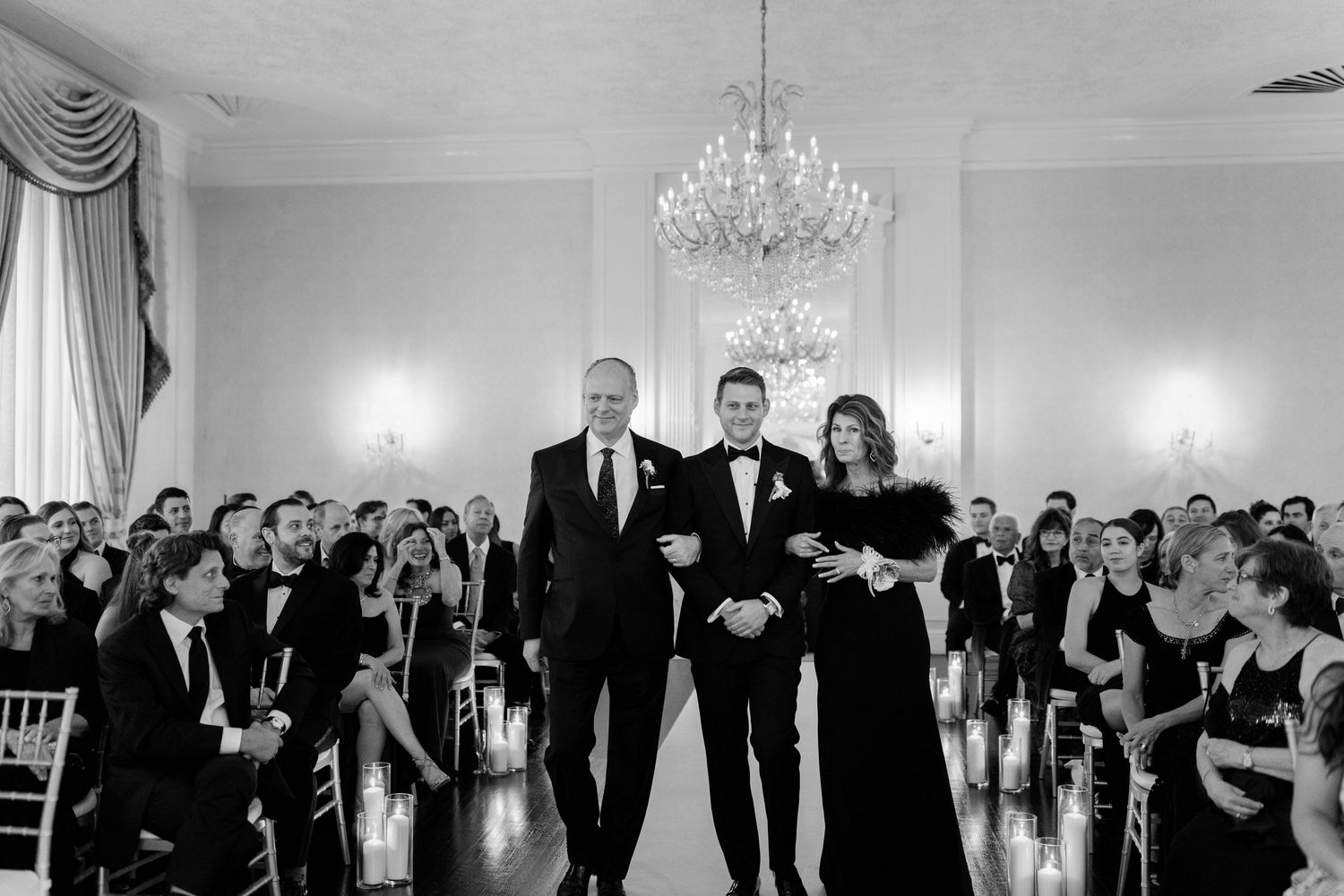 A formal ceremony takes place in an elegant ballroom with a crystal chandelier and rows of seated guests.