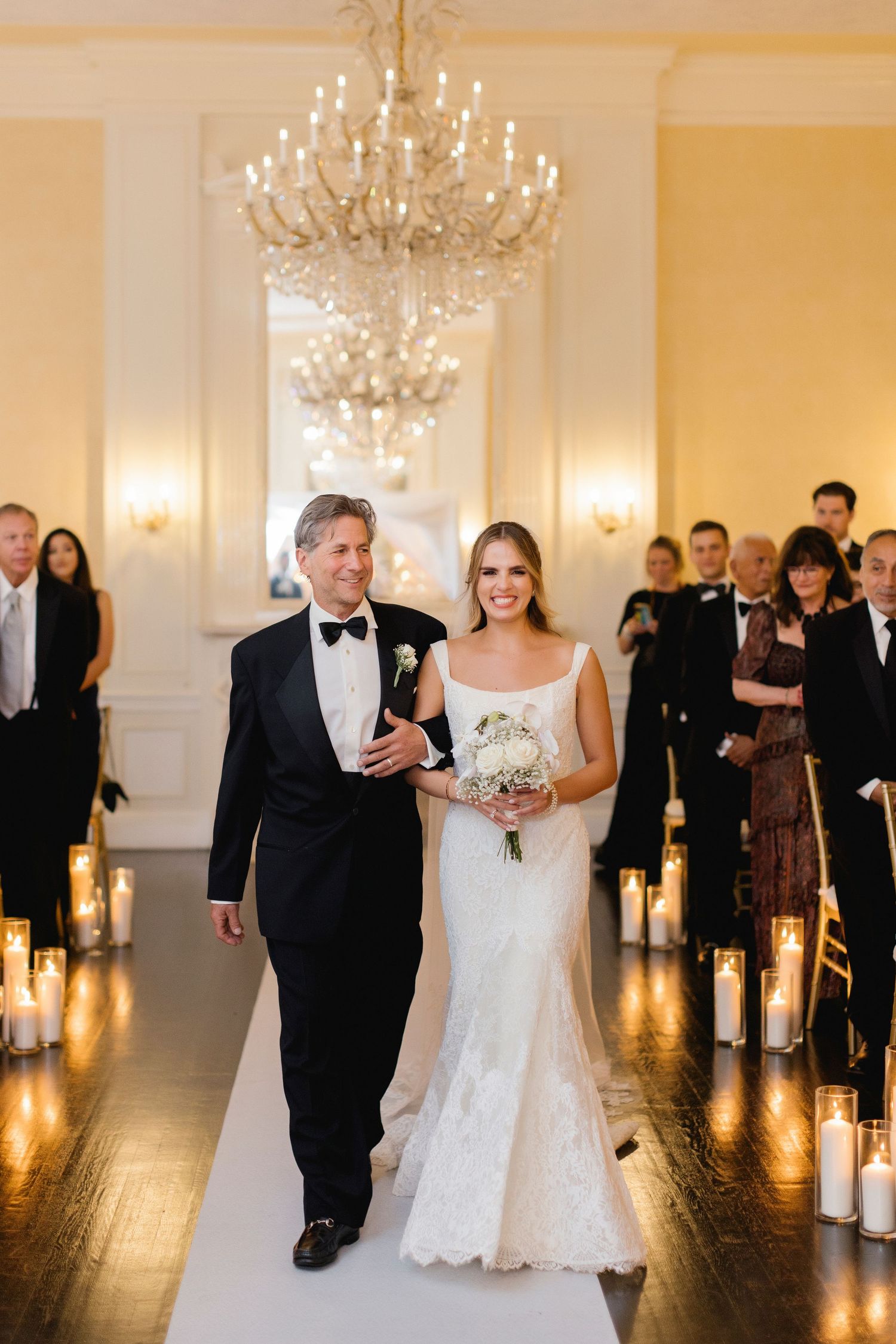 Wedding processional in elegant ballroom with chandelier and candlelit aisle.