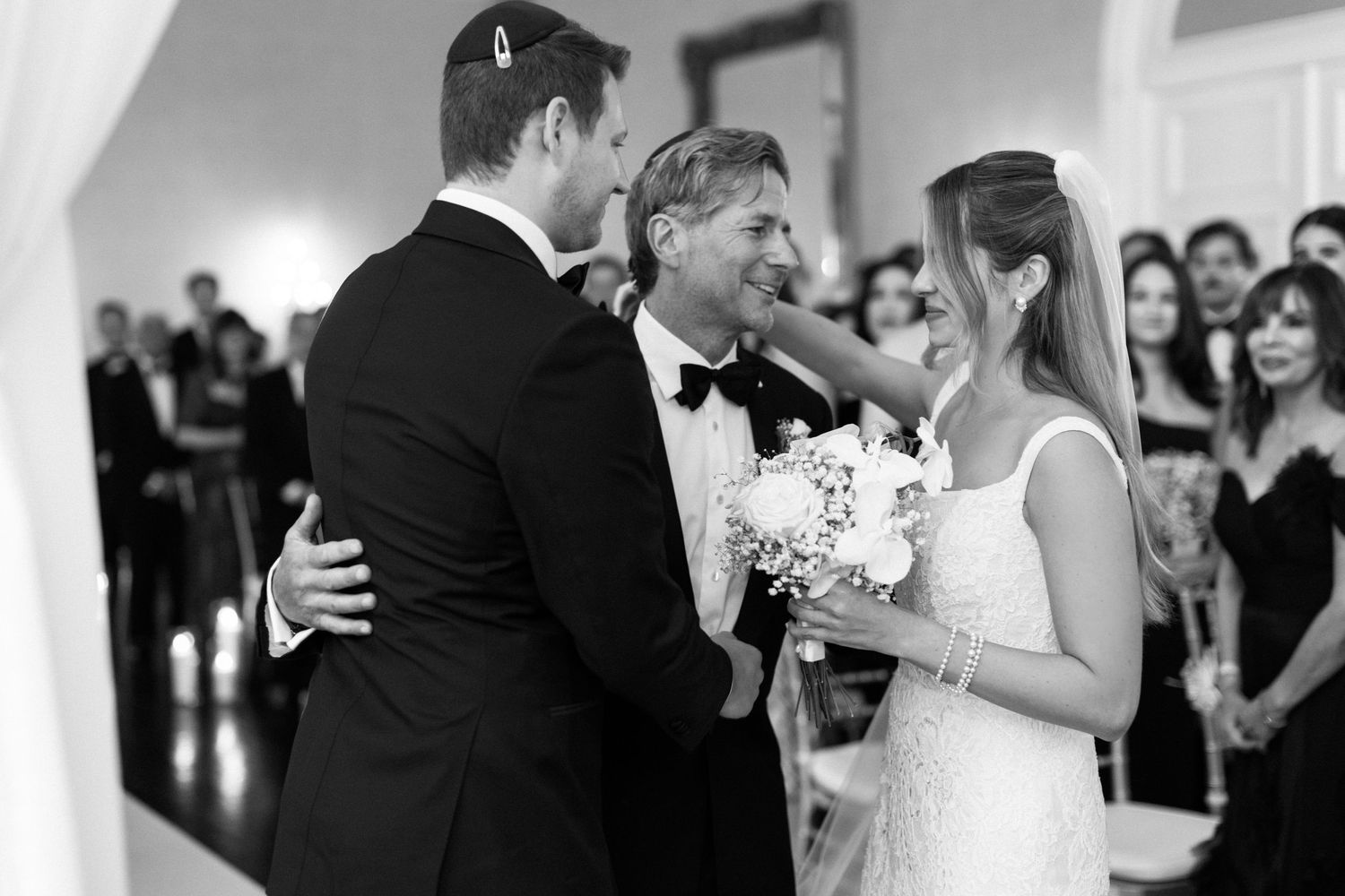 A black and white photo of a couple in wedding attire with guests watching in the background of an elegant venue.