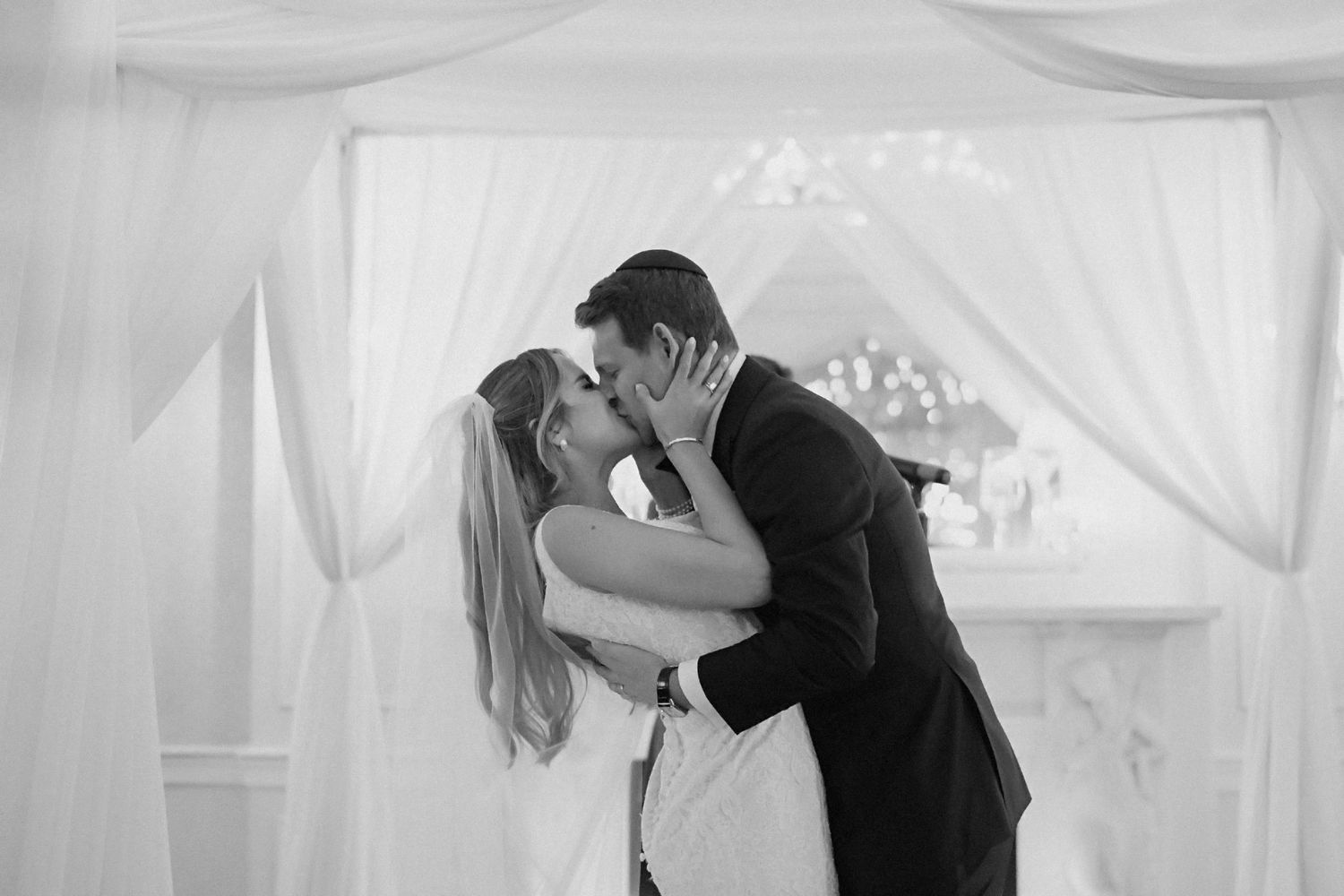 Black and white photo of newlyweds sharing romantic kiss under white fabric draping at wedding ceremony.