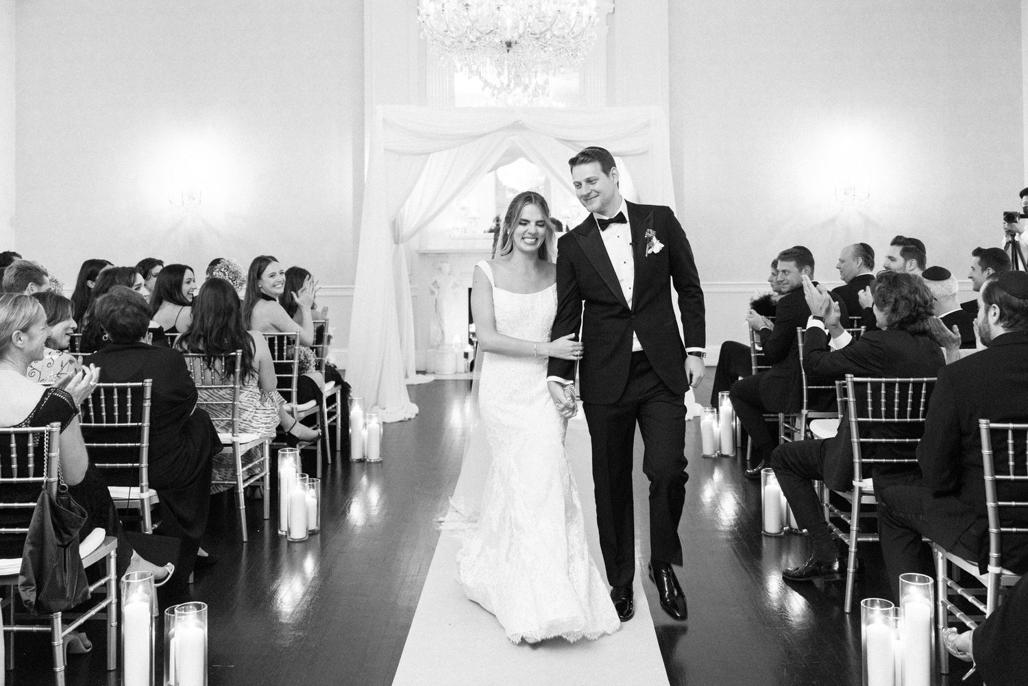 Newlyweds walk down the aisle together in a bright ceremony space lined with candles.