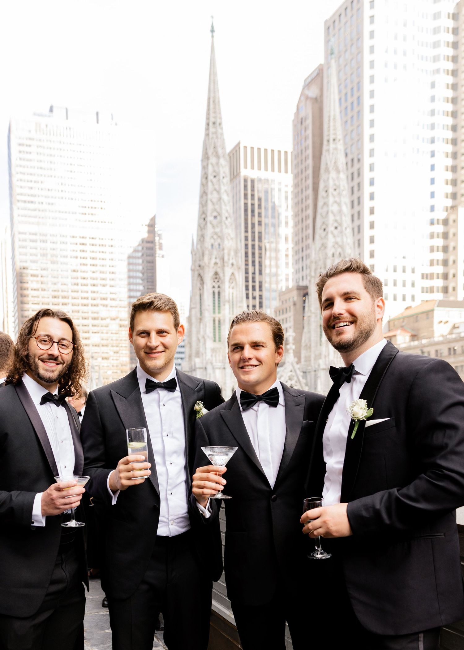 Four groomsmen in tuxedos toast with martini glasses against NYC skyline with St Patrick's Cathedral backdrop.
