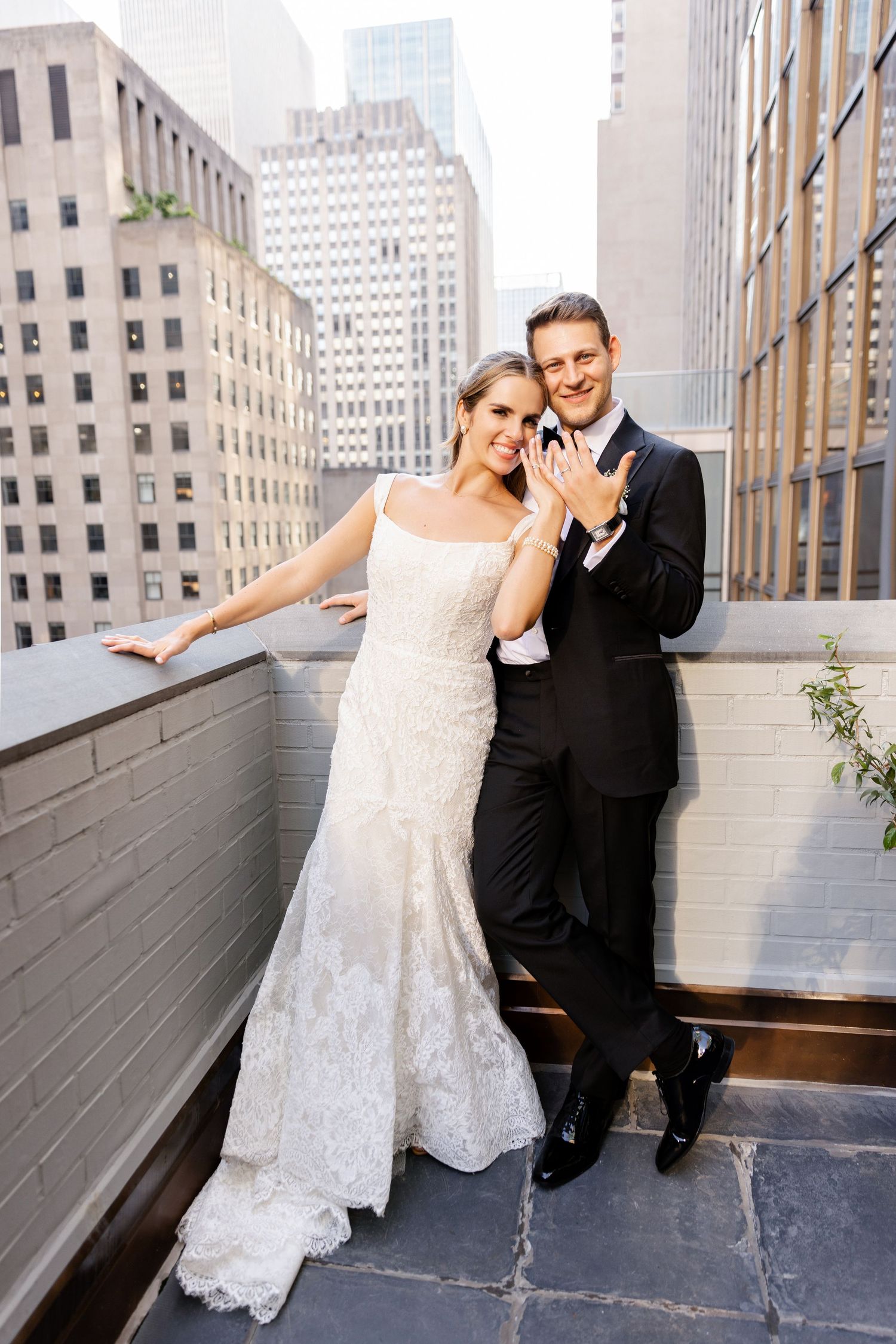 Couple in wedding attire poses on urban rooftop with city skyline backdrop.