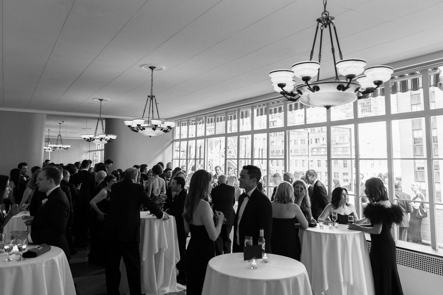 Black and white reception scene with guests mingling around cocktail tables near large windows.