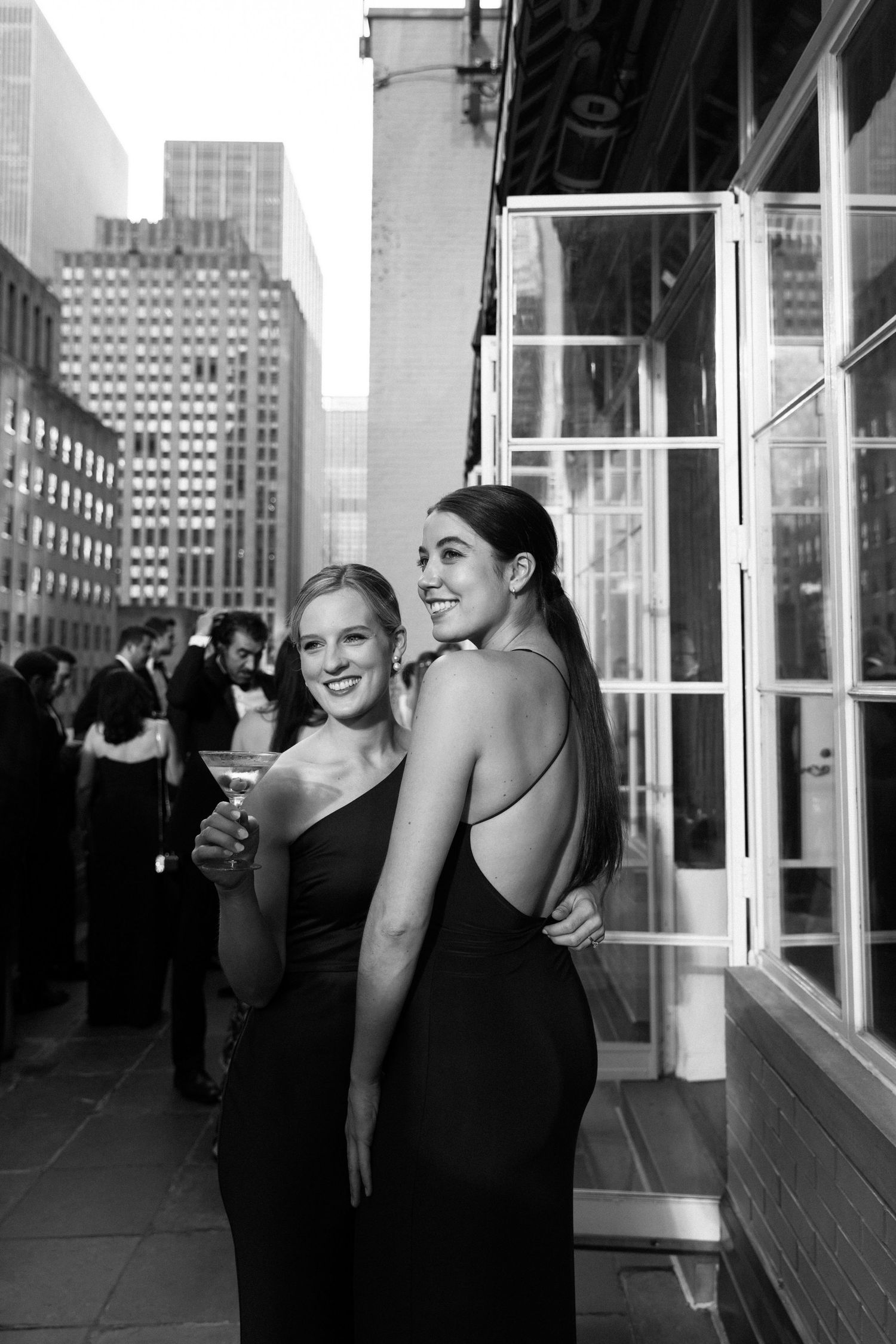 Two women in black formal dresses smiling on urban rooftop with city skyline in background.