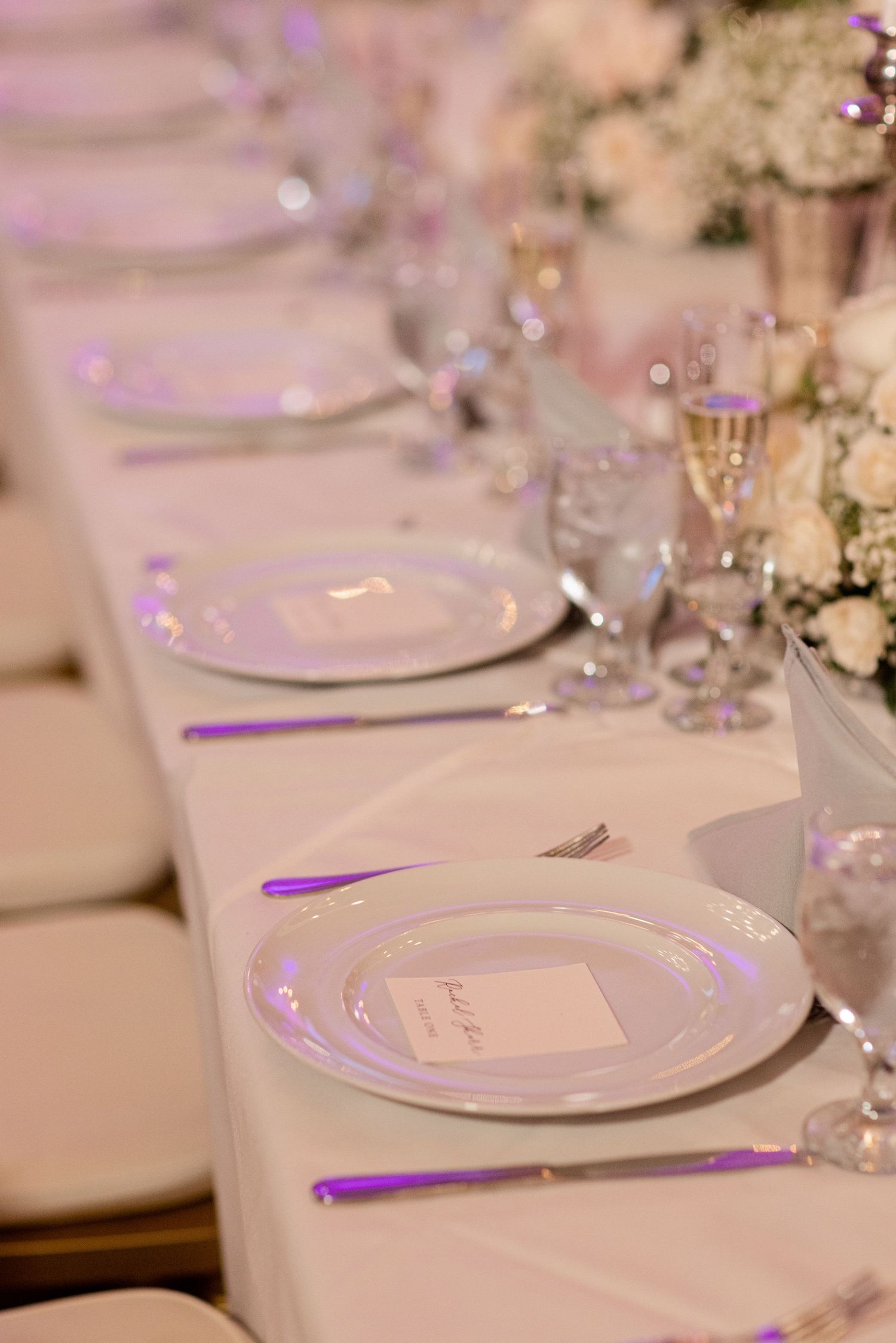 Wedding reception table setting with white plates and purple accents under soft lighting.