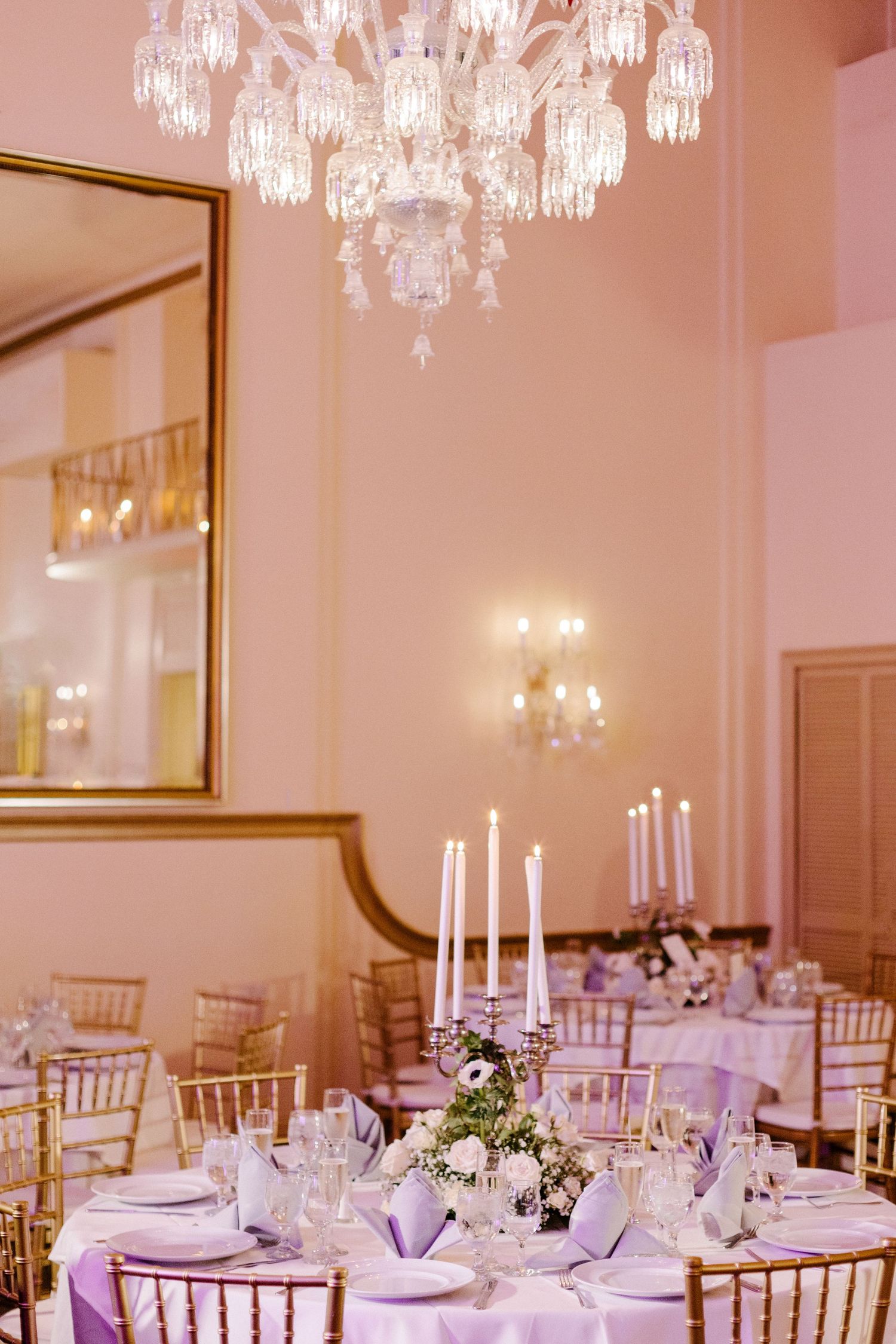 Luxurious reception room with crystal chandeliers and elegant table settings in soft pink lighting.