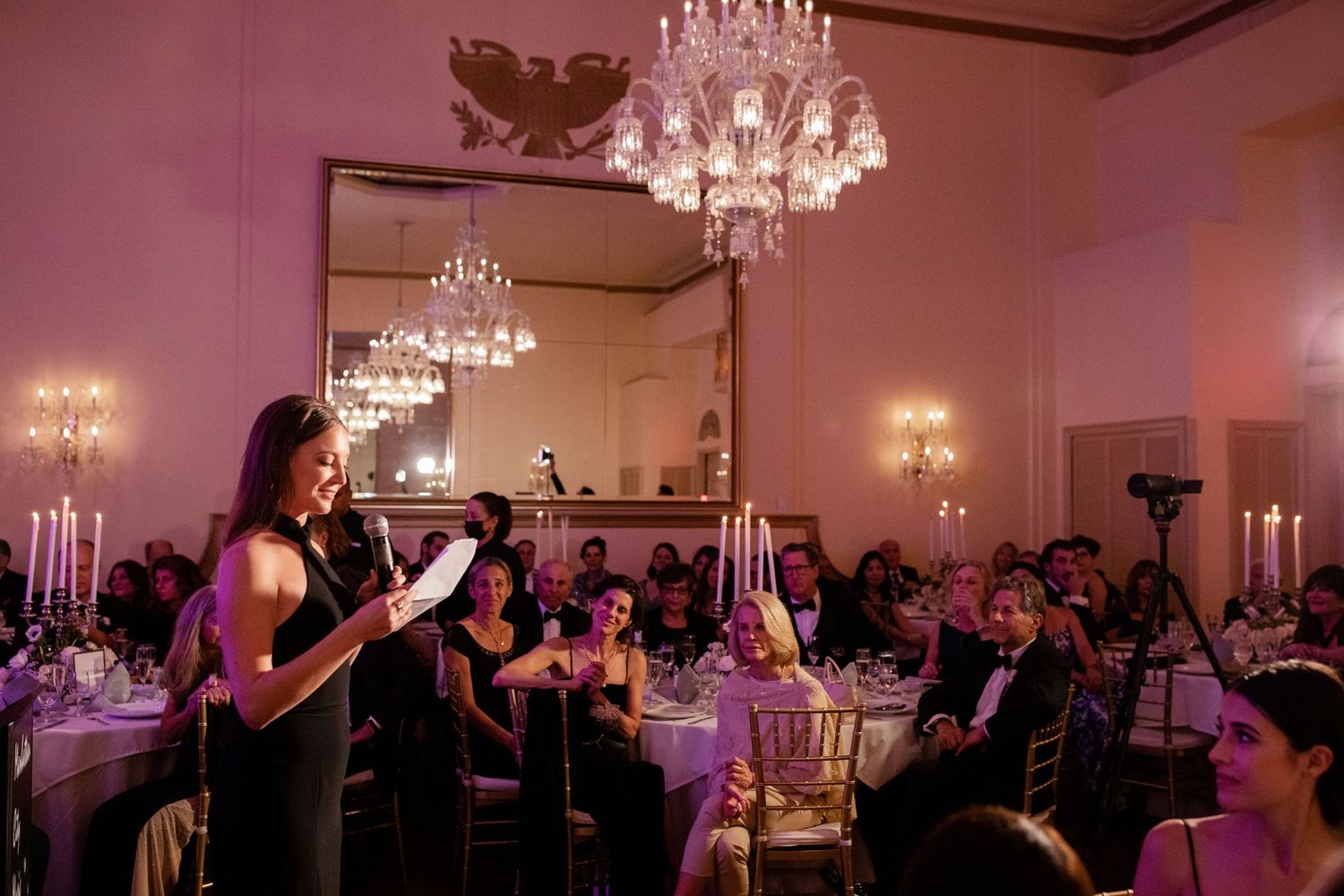 An elegant ballroom event with crystal chandeliers, pink ambient lighting and guests at round tables watching a performer.