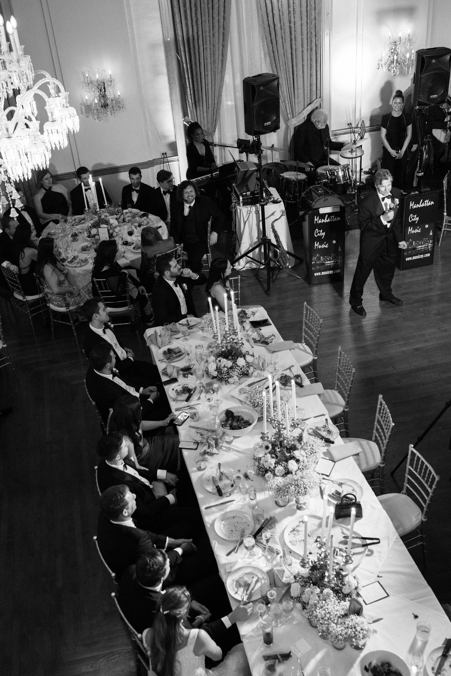 Black and white photo of wedding reception with live band performing and guests seated at long tables.