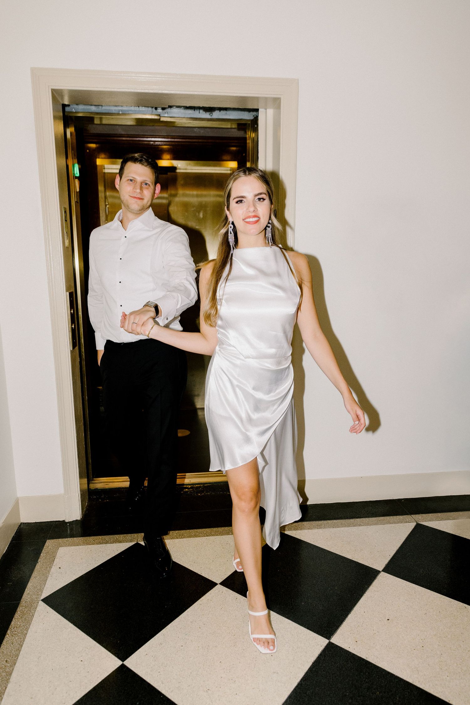 Couple in elegant attire poses on a black and white checkered floor near a doorway.