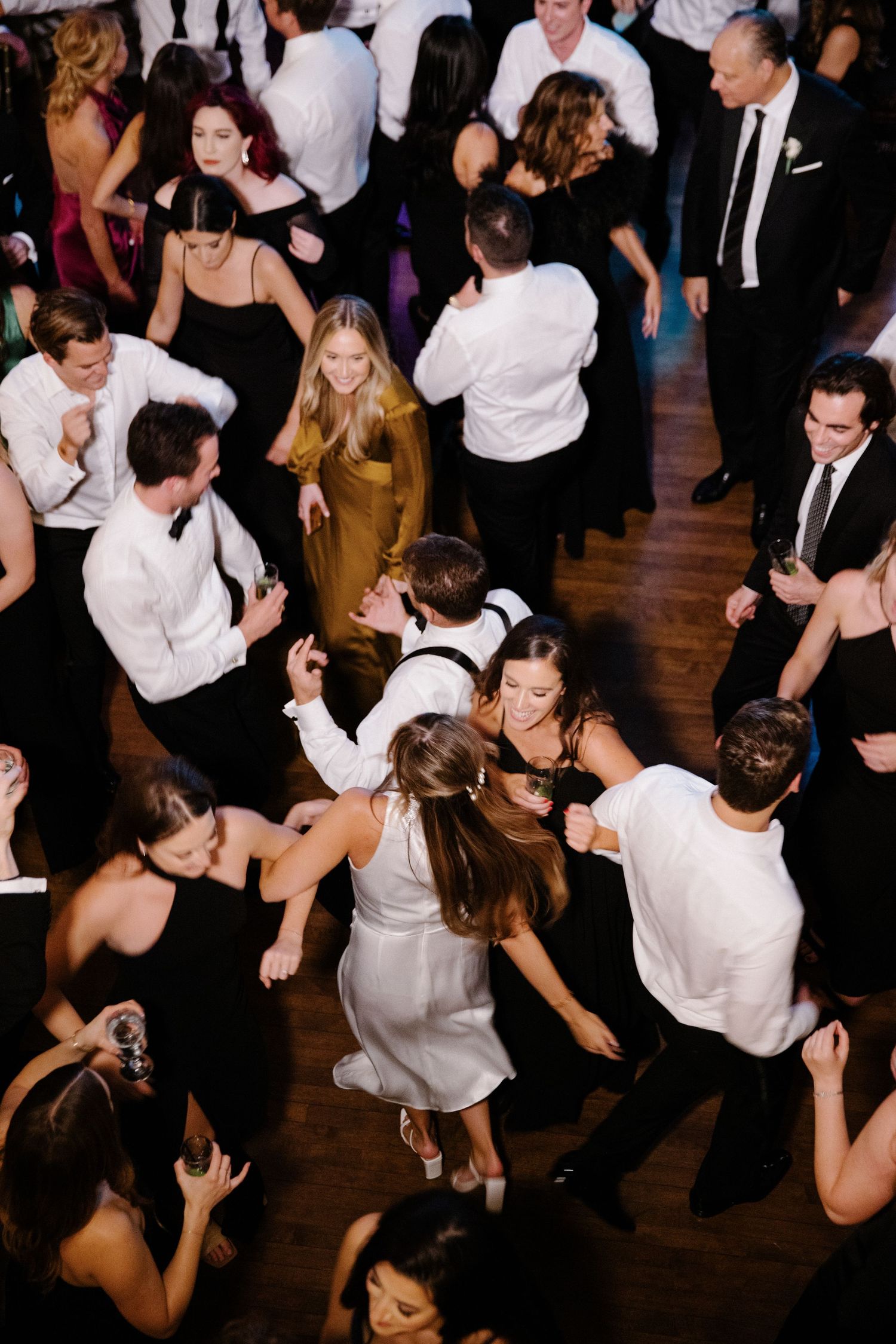 Crowded dance floor at a formal event with people dancing and mingling in formal attire on a wooden floor.