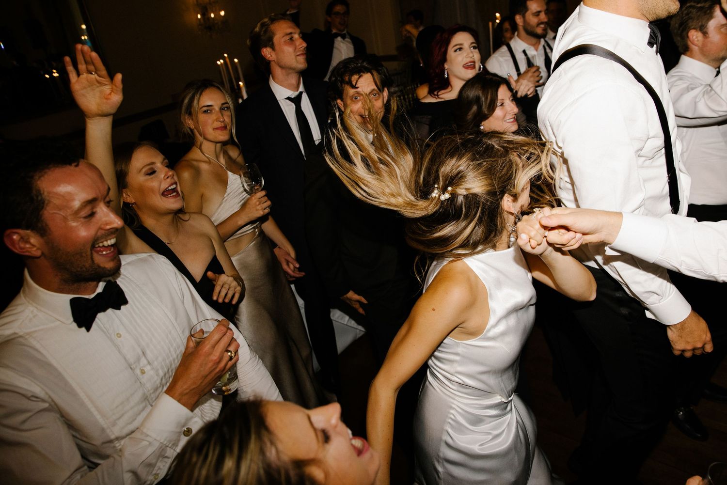 A crowd of wedding guests dance and celebrate energetically on a dimly lit dance floor at a reception party.