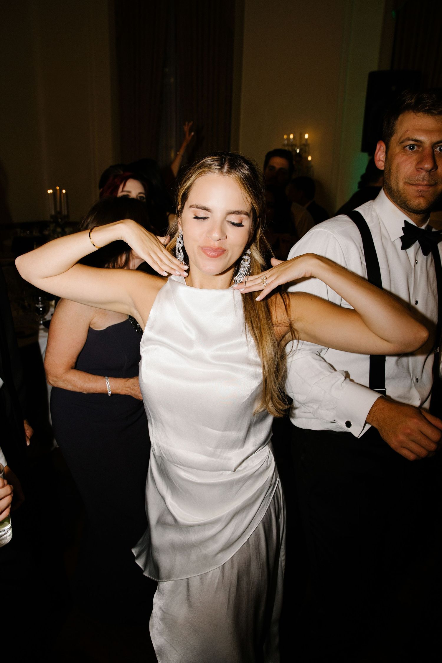 Person in a white satin dress dances joyfully at a wedding reception while others celebrate in formal attire.