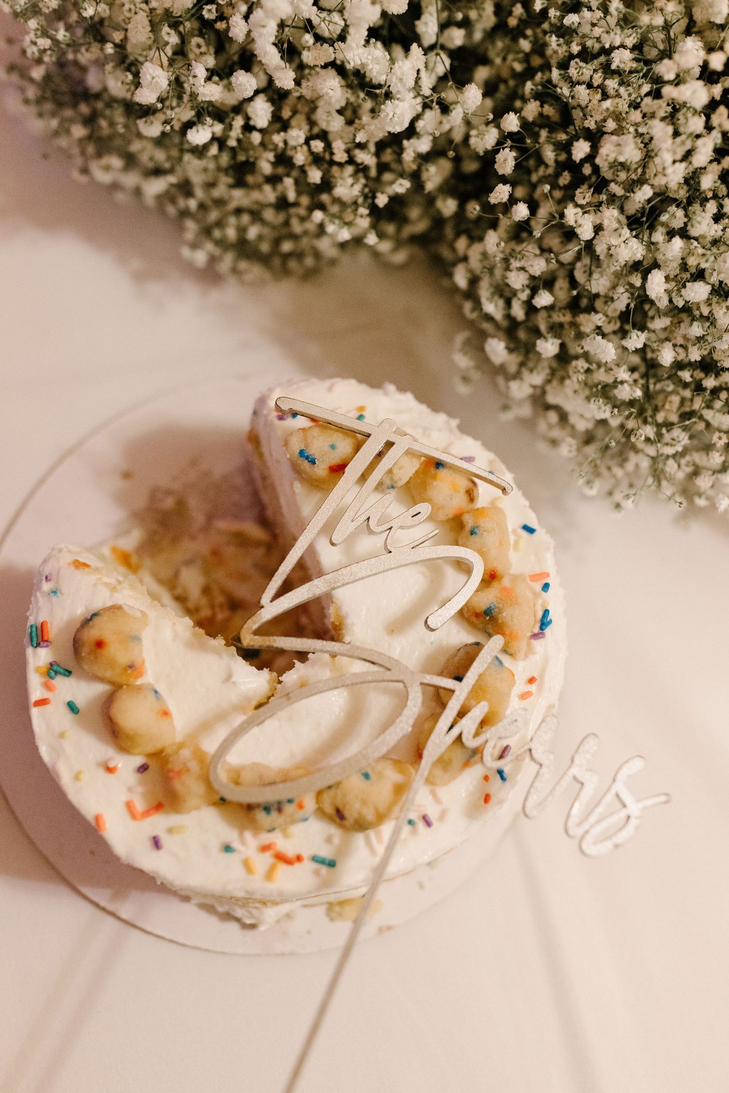 A decorative white frosted cookie or treat with colorful sprinkles sits on a plate near baby's breath flowers.