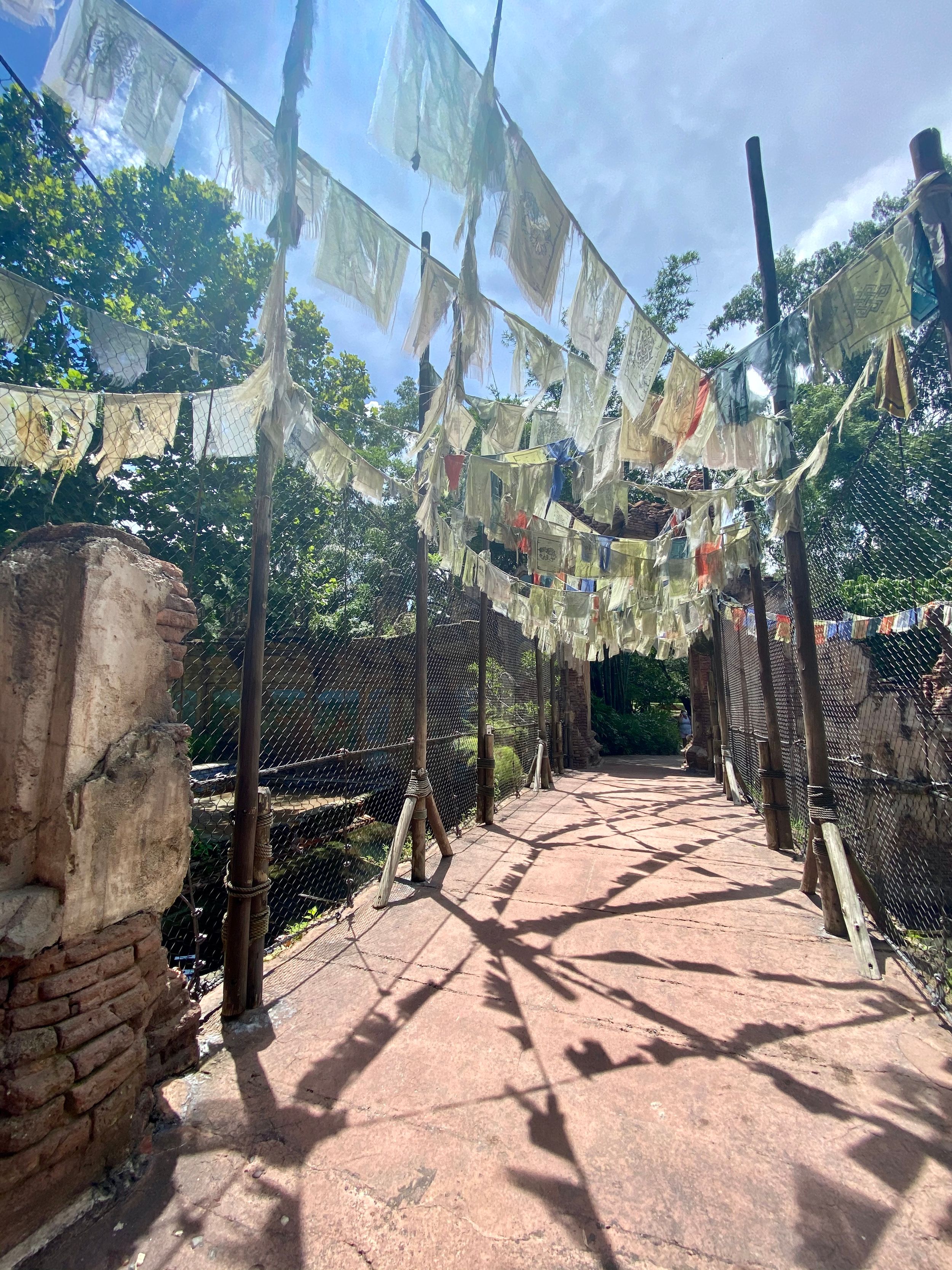 Tibetan prayer flags hang over a winding stone pathway under blue sky, creating shadows on the pink-tinted walkway below.