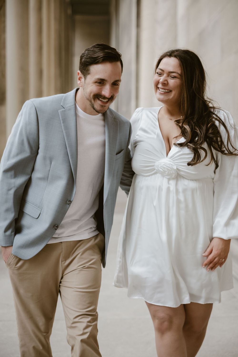 A romantic couple shares a candid moment together, with him in a gray blazer and her in a white dress against neutral walls.