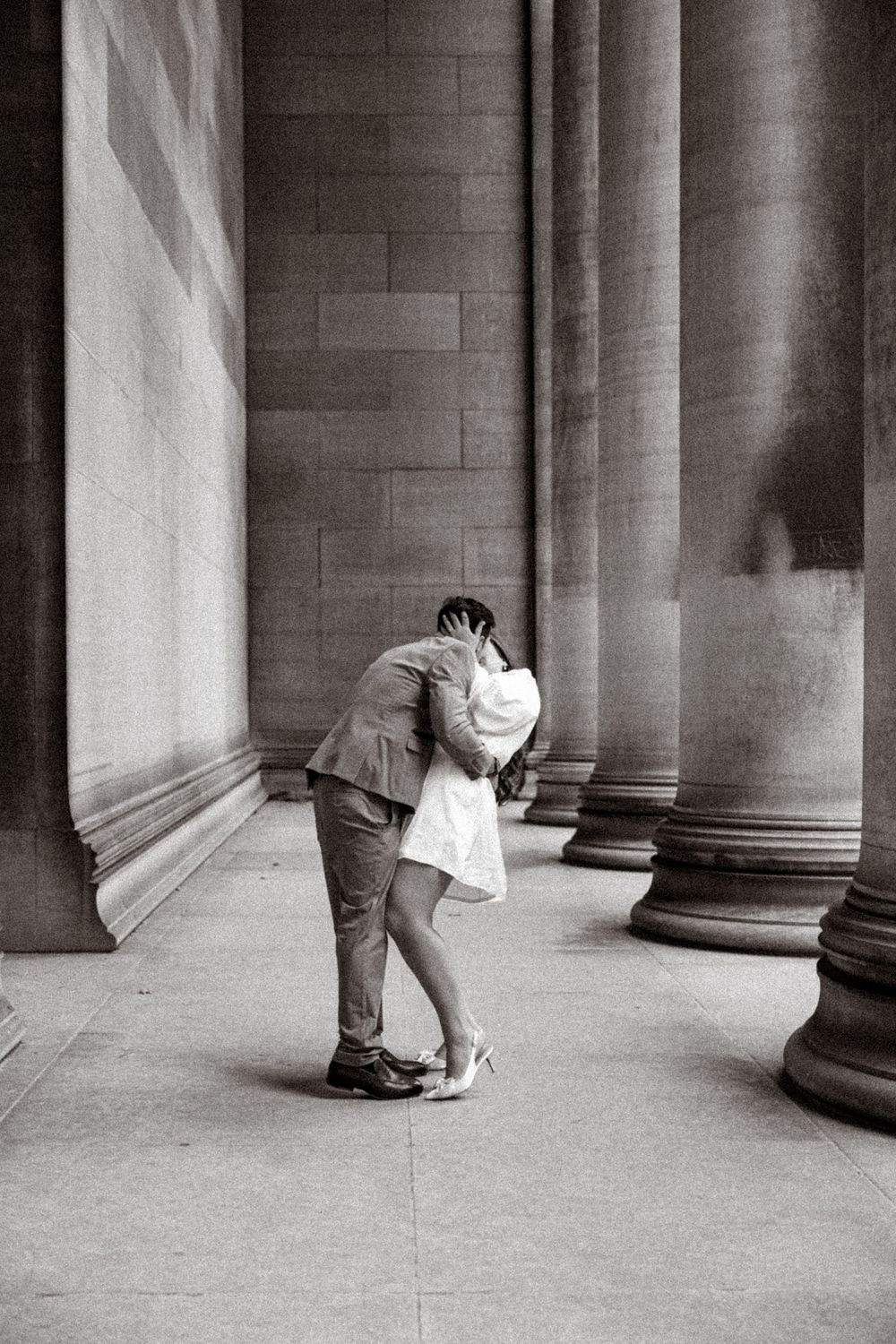 A romantic black and white photograph of a couple embracing between classical stone columns.