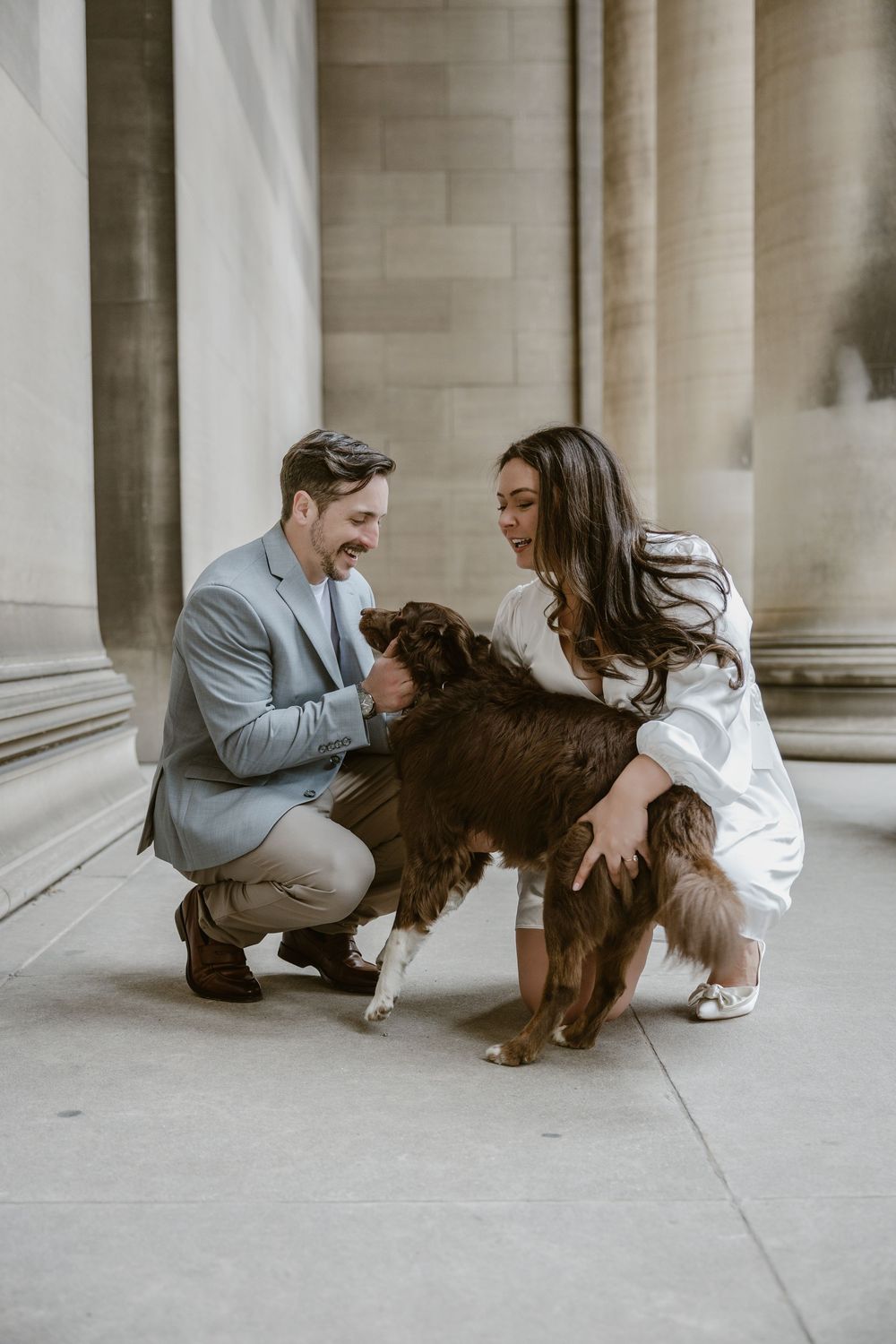 A couple kneels next to their brown fluffy dog on the sidewalk in front of a building with stone columns.