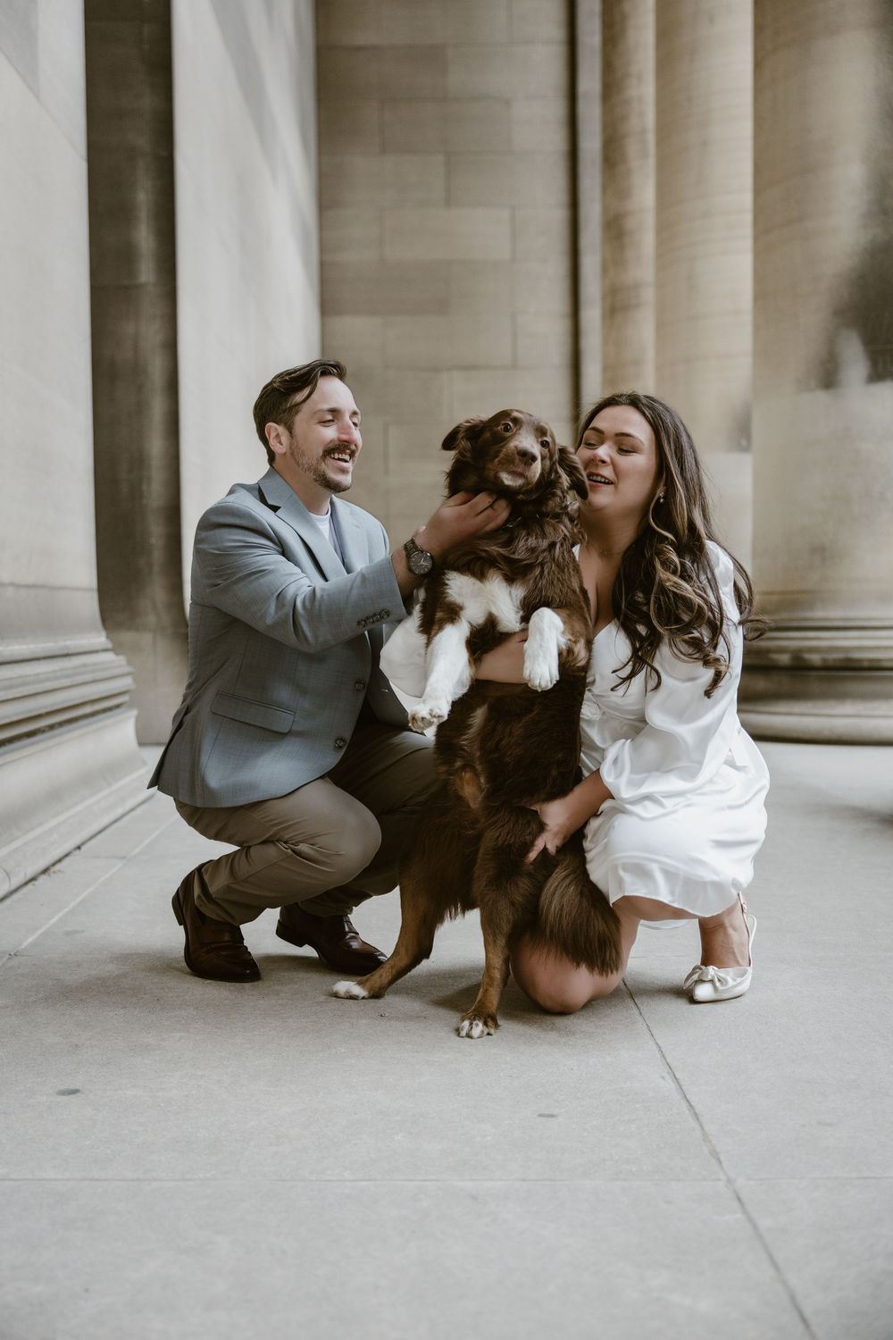 Couple wearing grey suit and white dress laughing with their brown dog between pillars at engagement photoshoot.