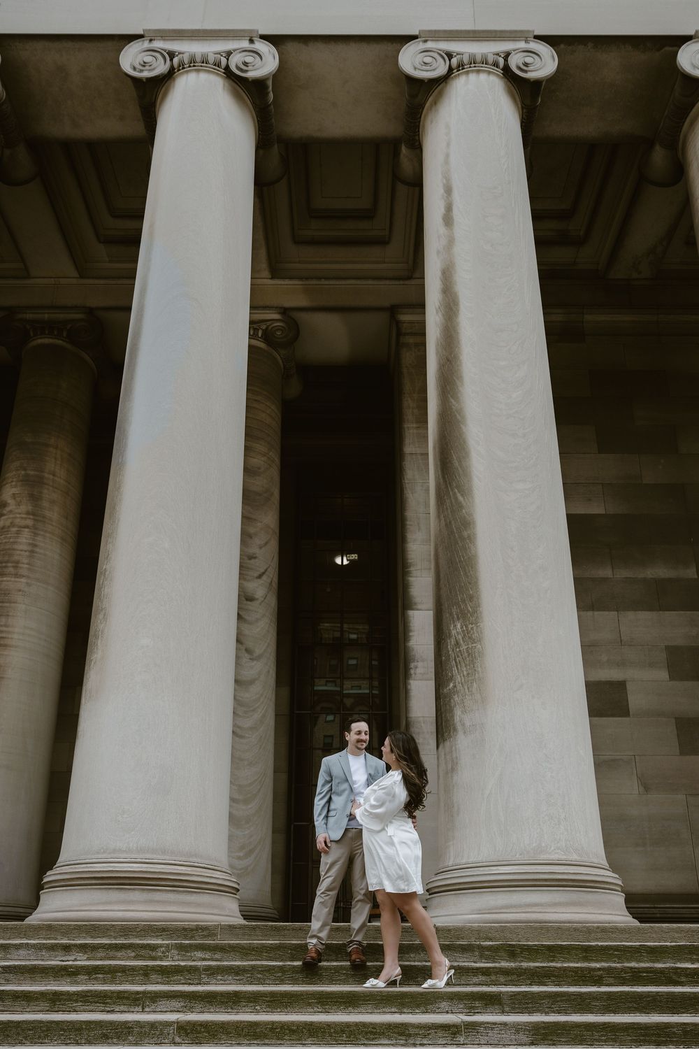 Two people stand between towering Ionic columns on stone steps of a neoclassical building.