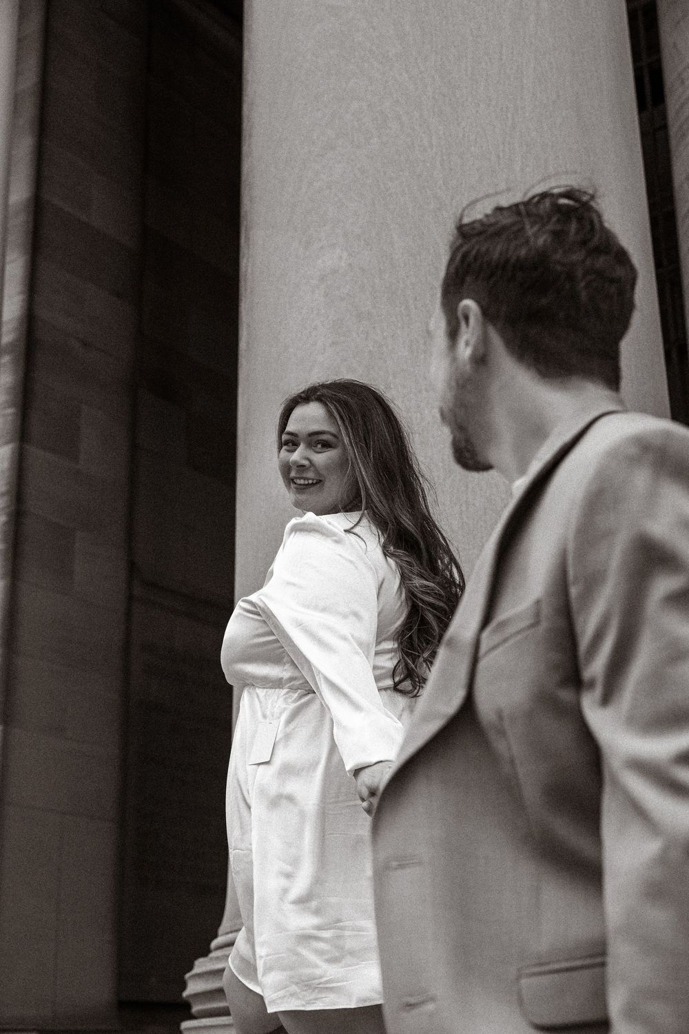 A woman in a white dress shirt smiles while standing near a column as a man in a suit looks on in black and white.
